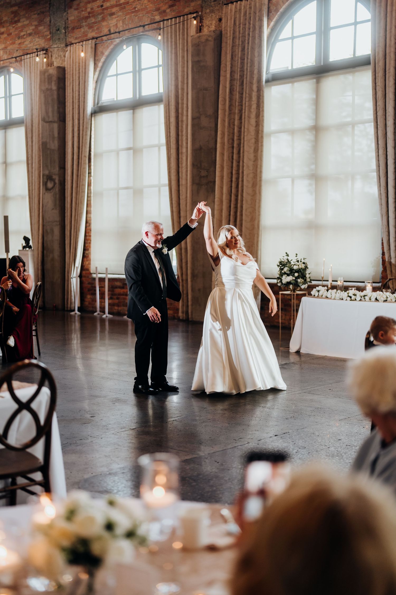 Wedding reception in an elegant ballroom with arched windows and candlelit tables.