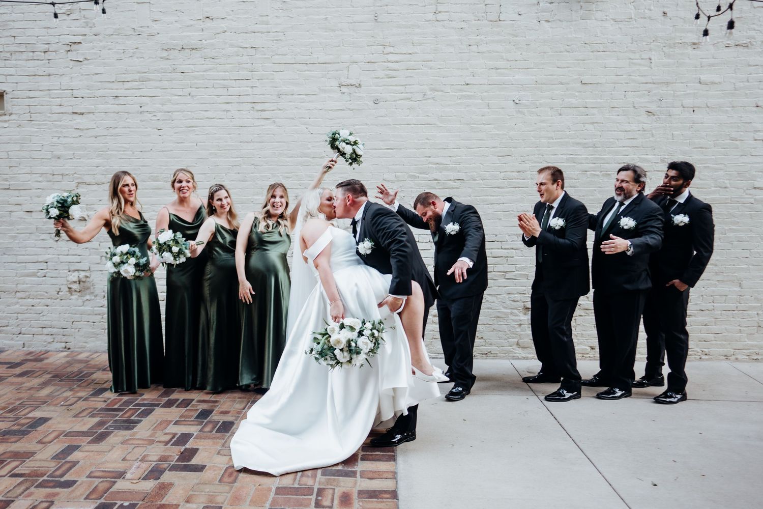 Wedding party members arranged in casual poses against a white brick wall background.