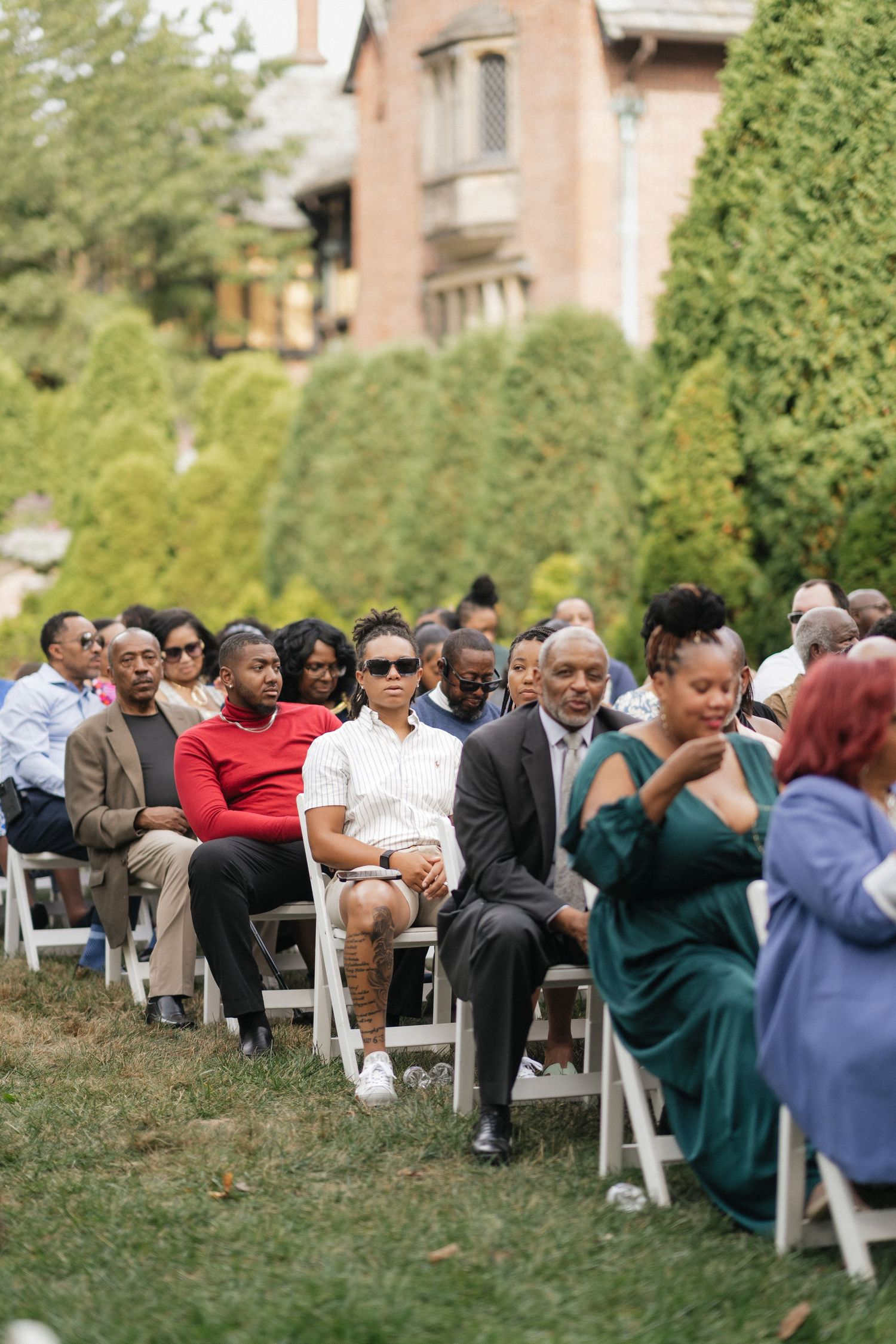 Wedding guests seated in white chairs during an outdoor ceremony with greenery in background.