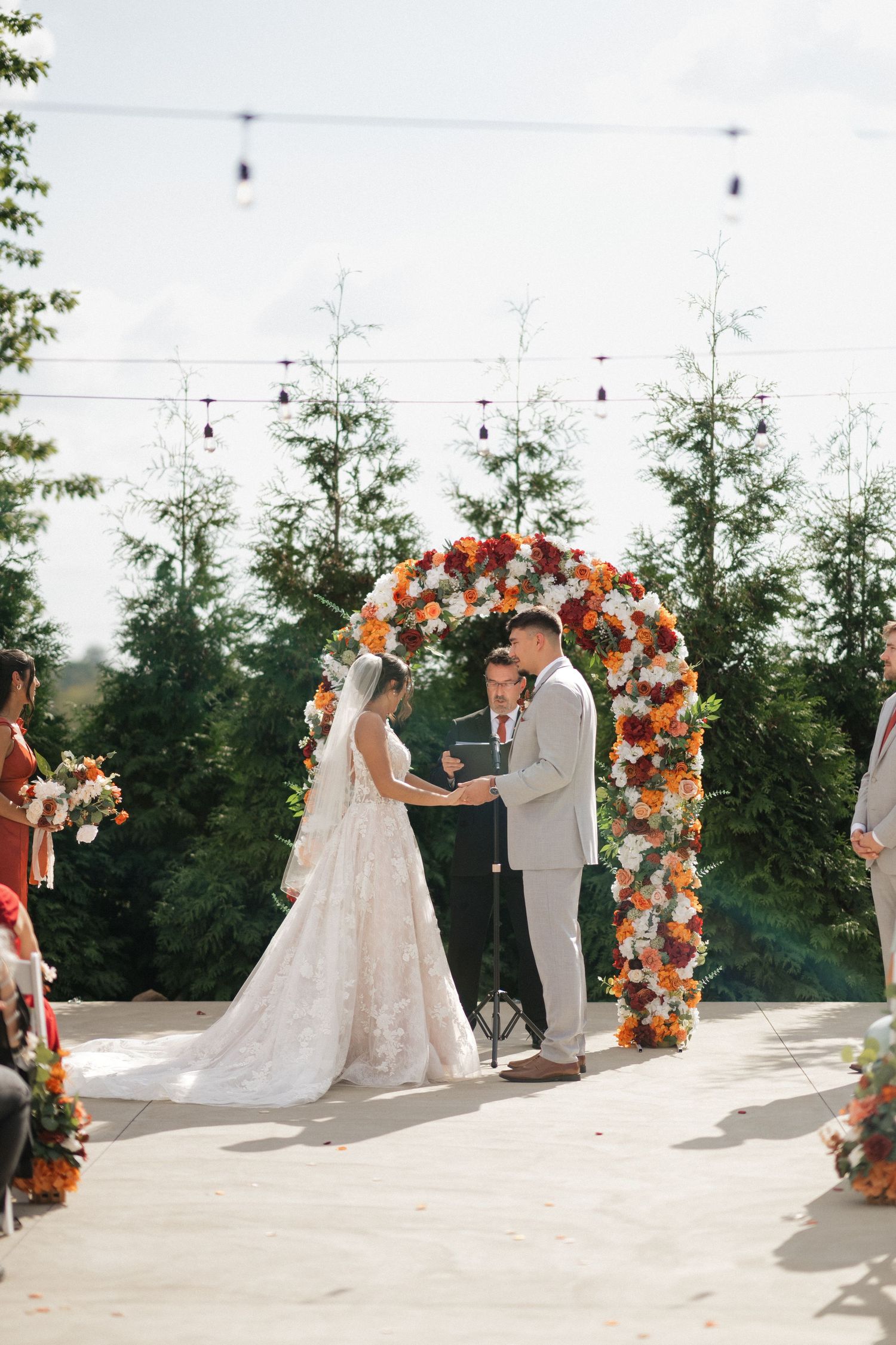 Outdoor wedding ceremony with coral and orange floral arch against evergreen trees.