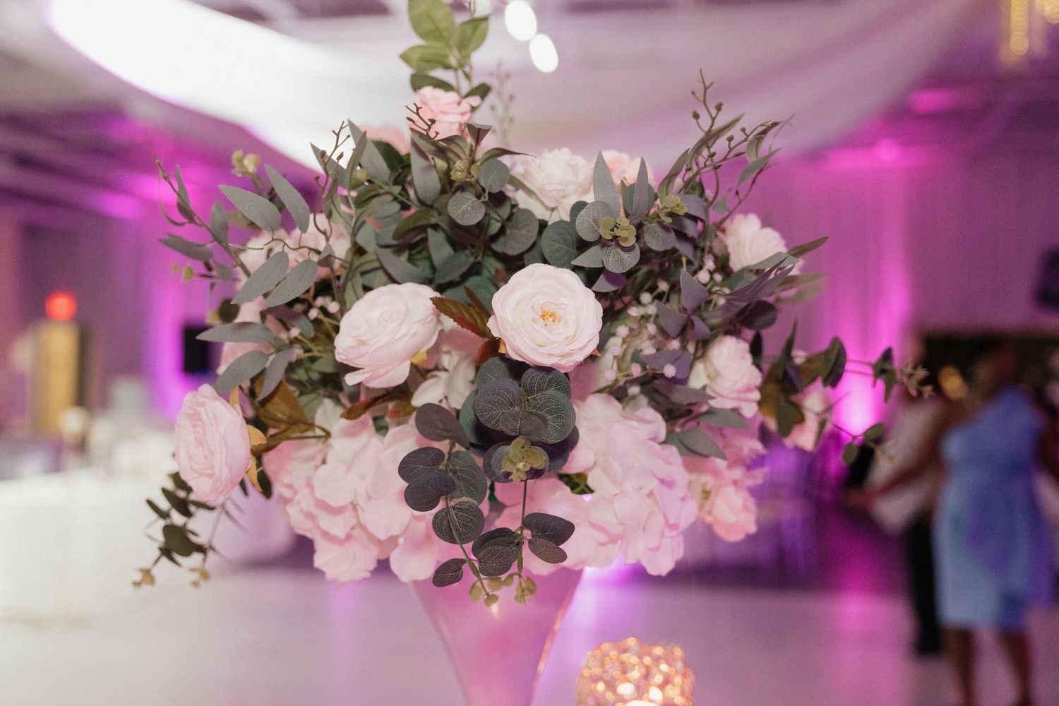 Pink and white floral centerpieces with eucalyptus under purple reception lighting.