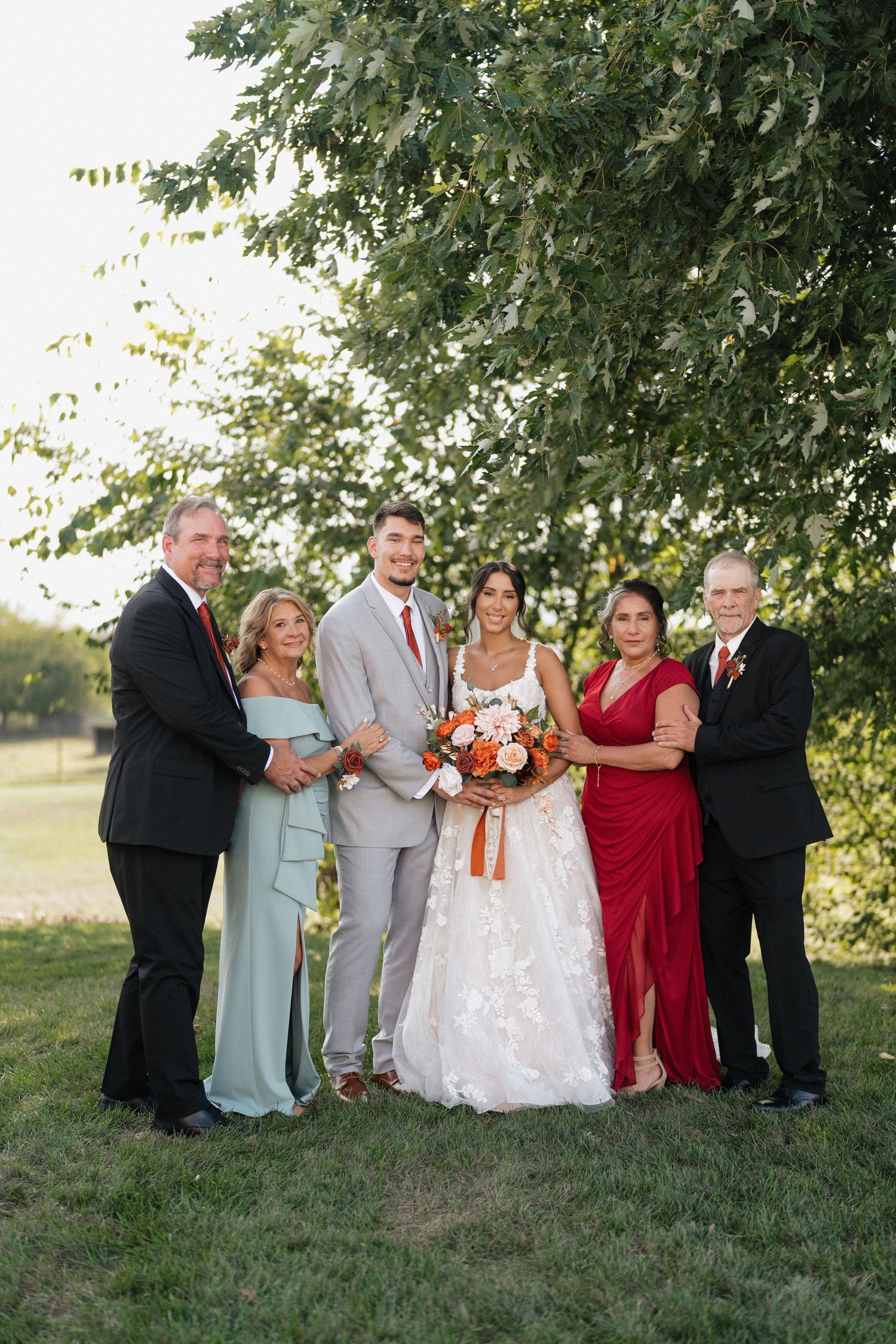 Wedding party poses together on grass under trees at sunset.
