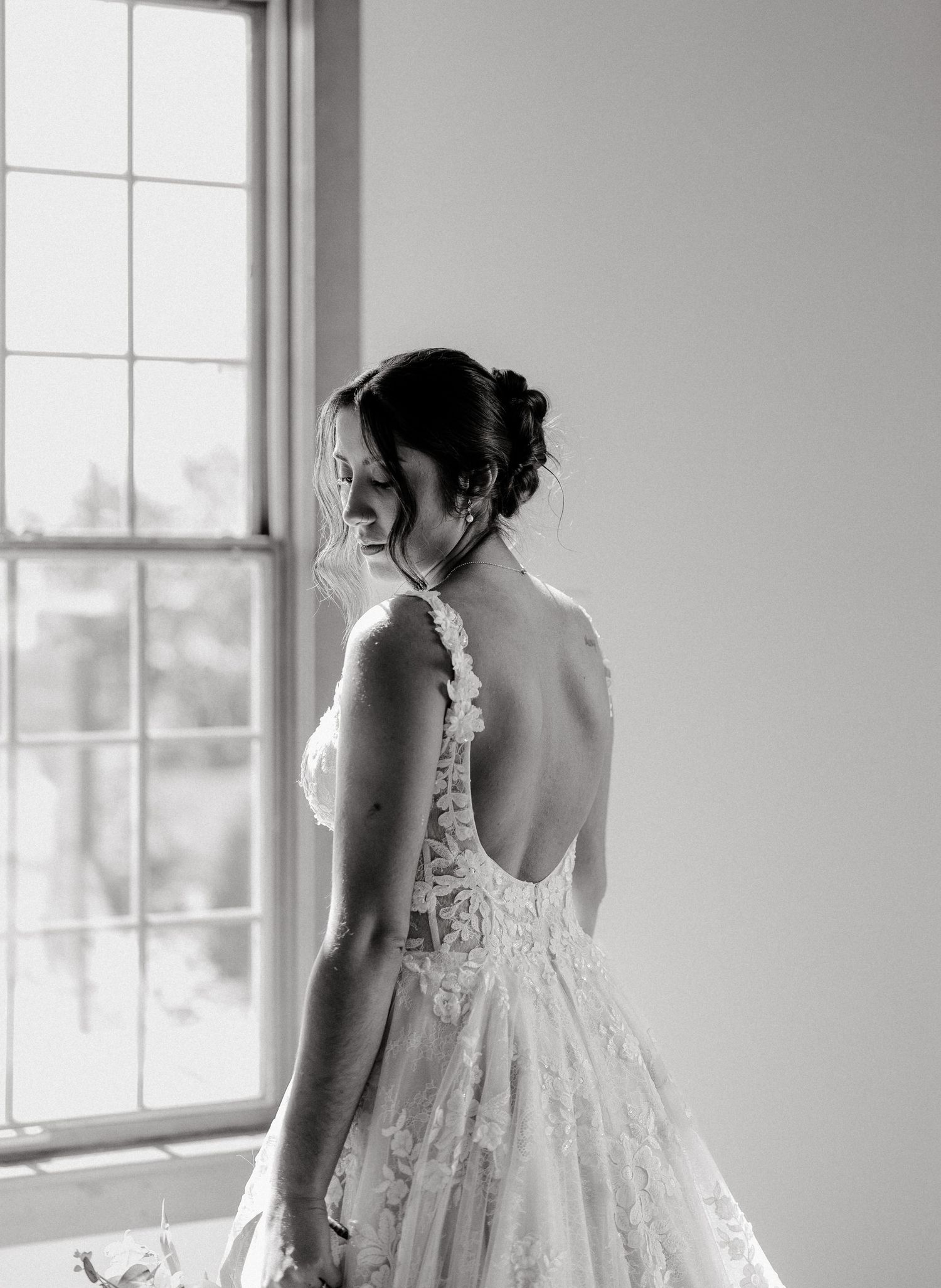 A bride in an elegant white lace dress poses near large windows in black and white.