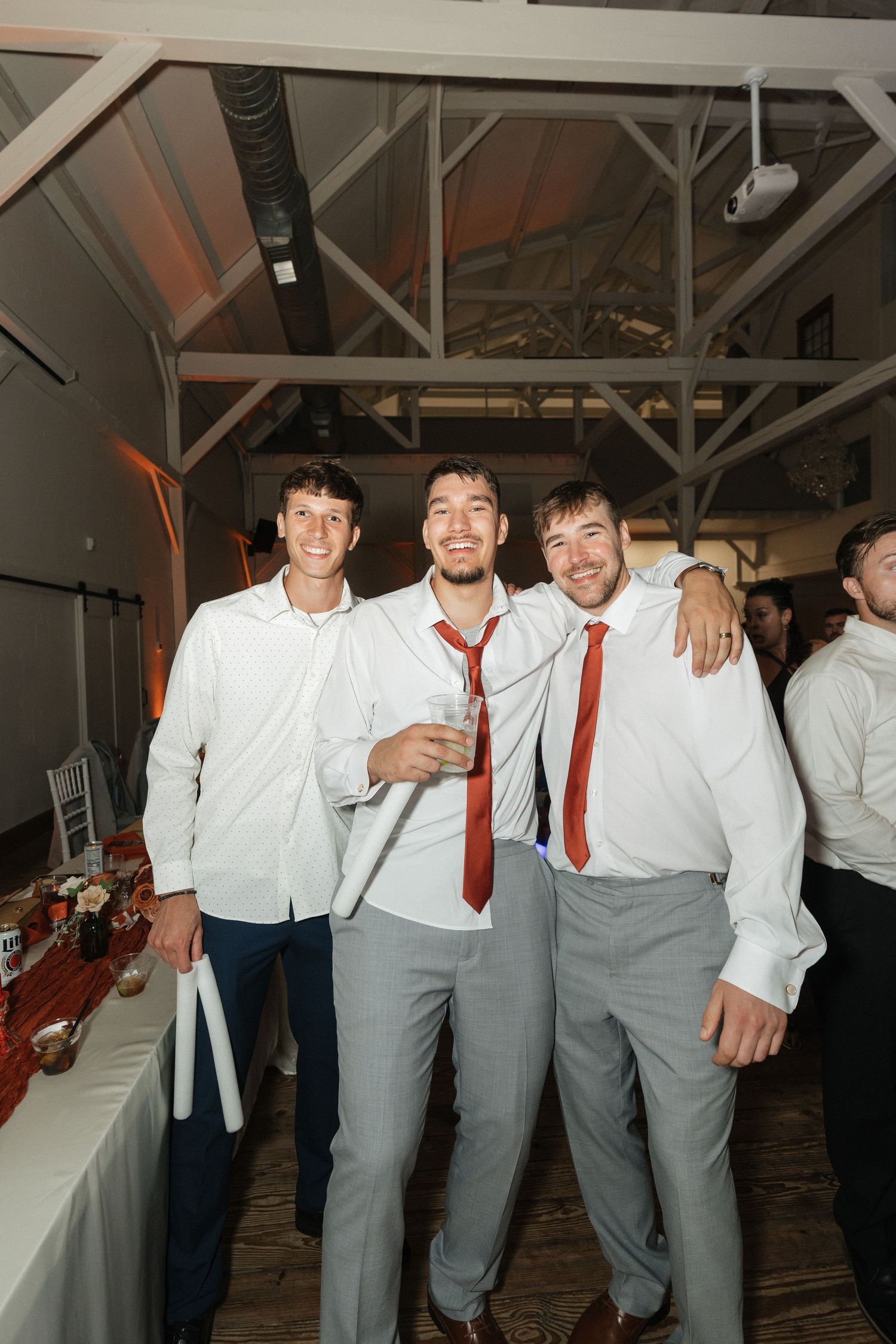 Groomsmen in white shirts and gray pants celebrate at indoor wedding reception.