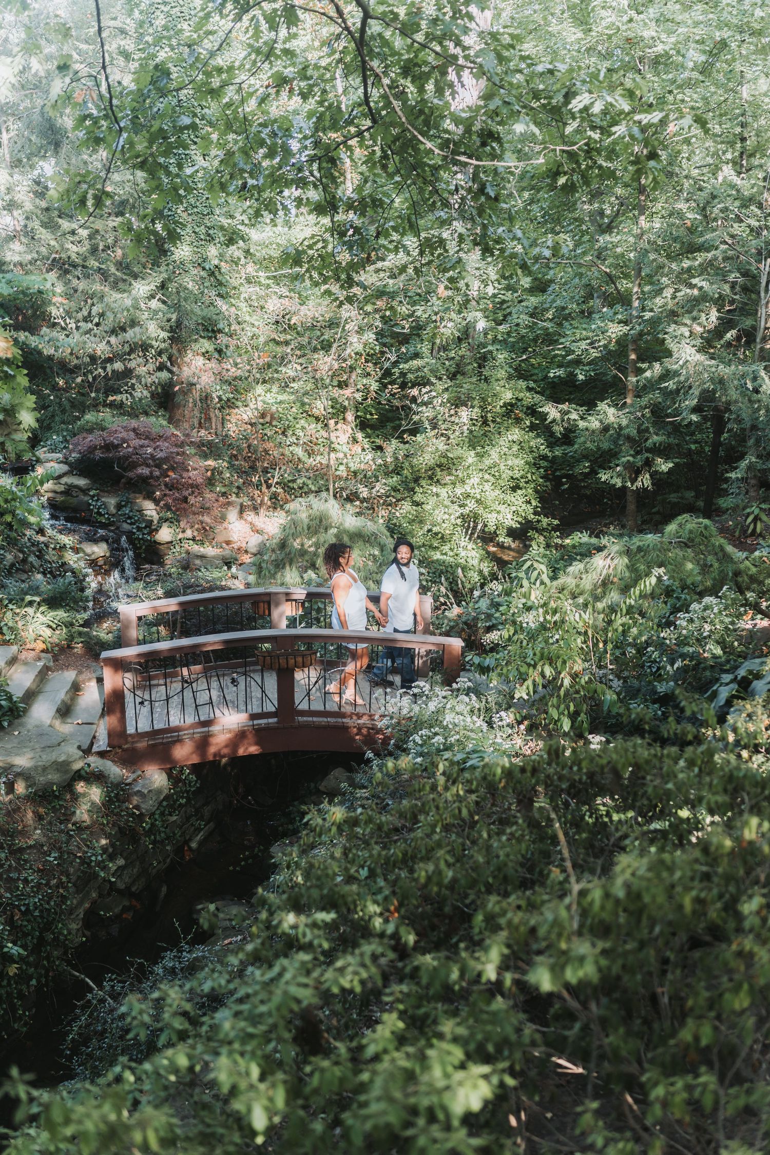 A couple stands together on a wooden bridge surrounded by lush green forest.