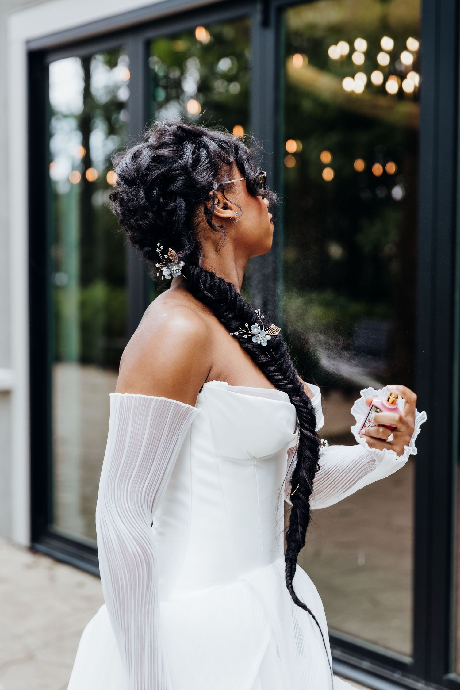Elegant bride in off-shoulder white dress with long braided hair adorned with flowers poses against window backdrop.
