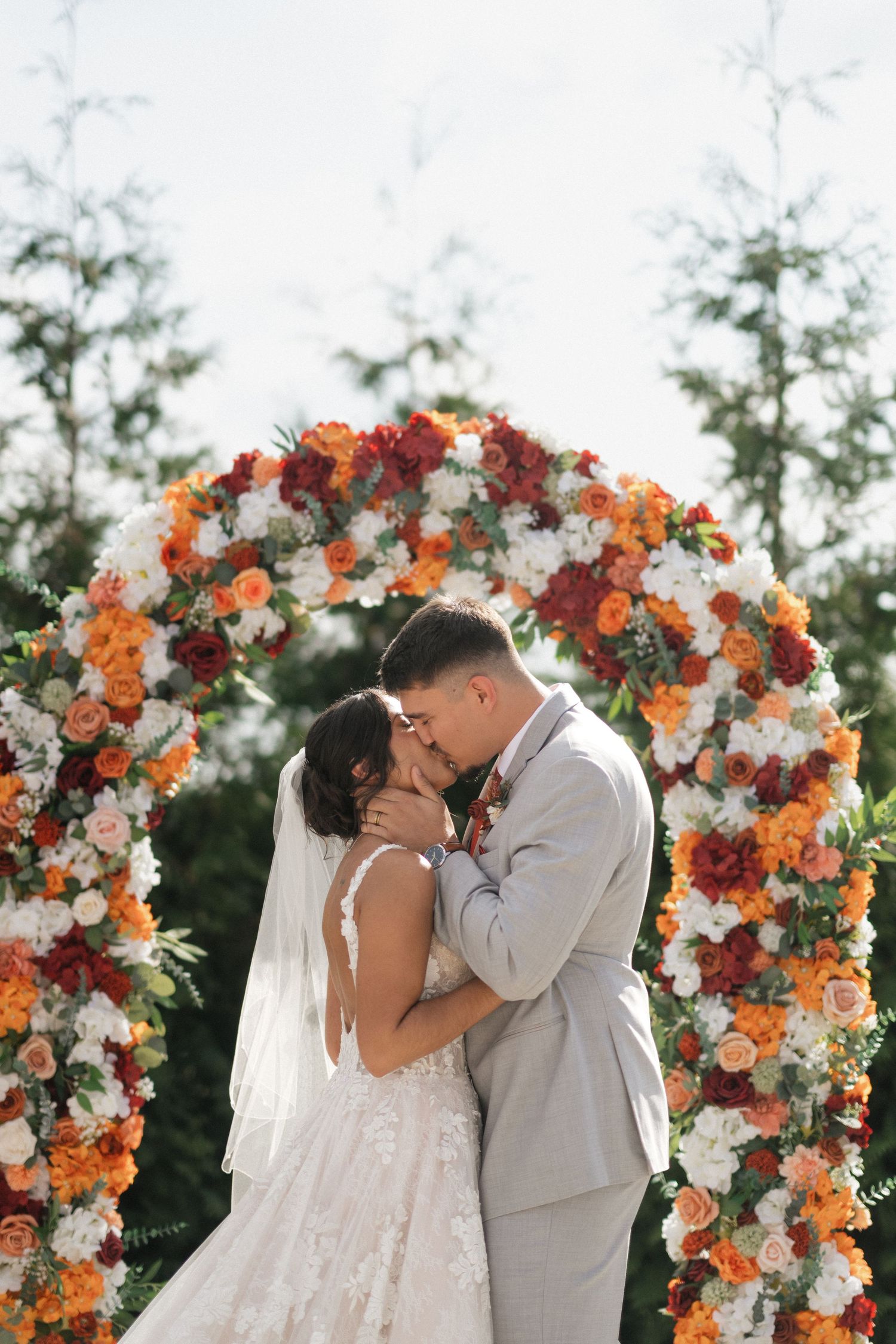 A couple shares their first kiss under an ornate arch of orange and white flowers at their outdoor wedding.