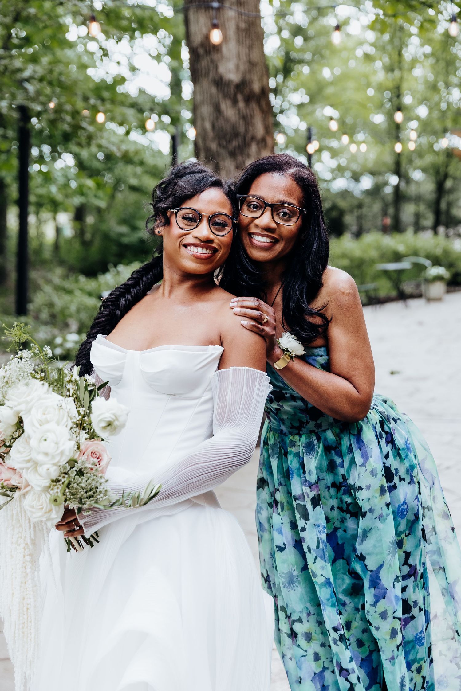 Wedding party members embrace and laugh while wearing white gown and blue floral dress outdoors.