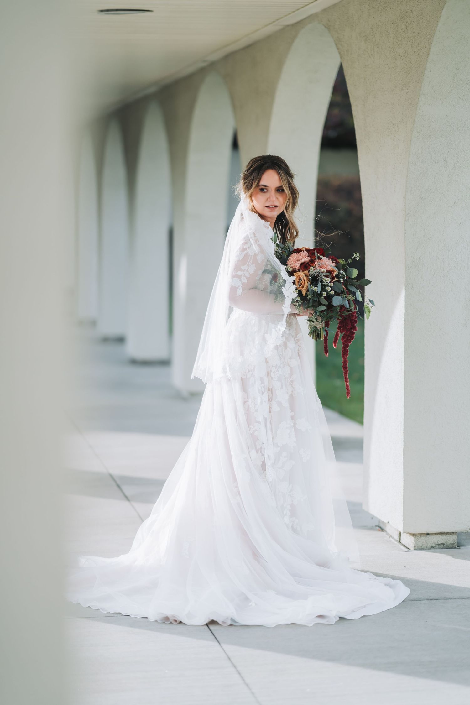 A bride in a flowing white lace wedding dress with a long veil poses gracefully in a white columned corridor.