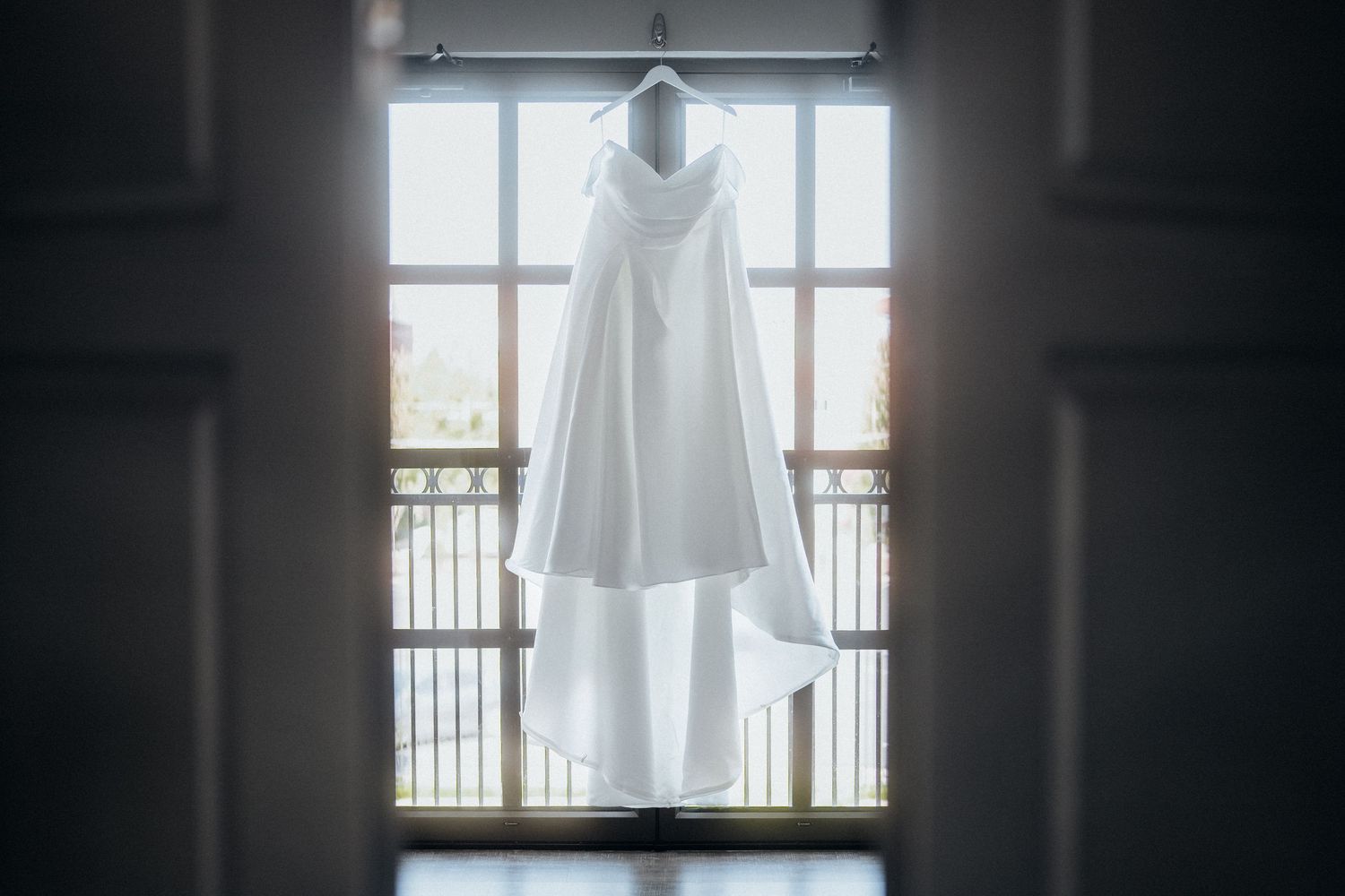 Wedding dress hanging in window creating dramatic silhouette against natural light.