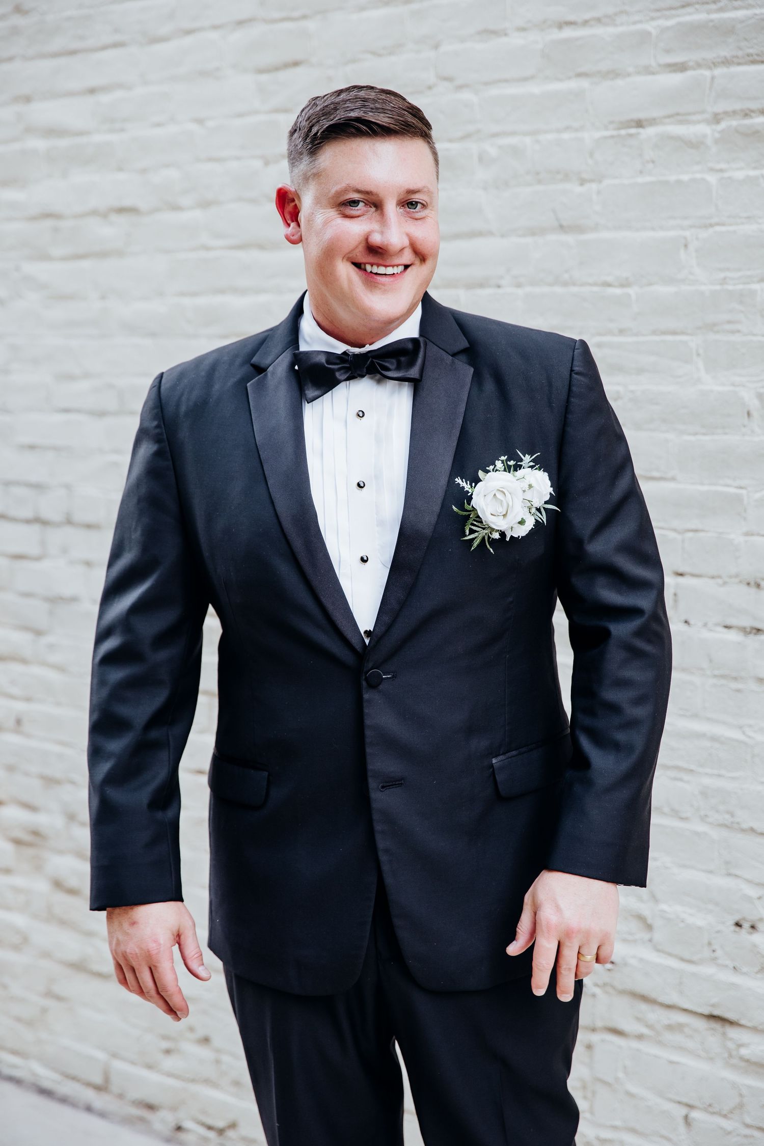 A groom in a classic black tuxedo poses against a light colored wall.