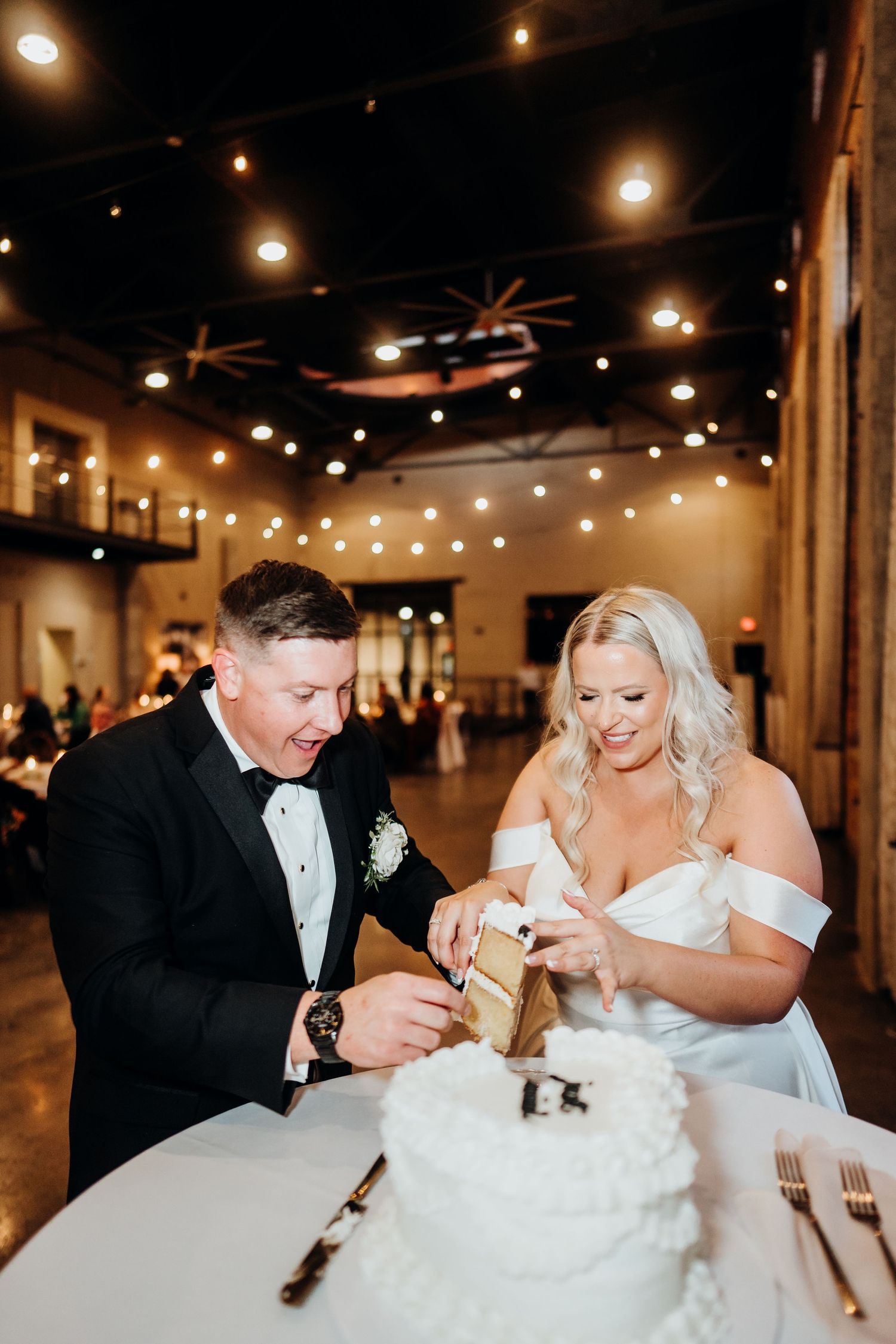 Newlyweds share a playful moment feeding each other wedding cake in a rustic venue with string lights overhead.