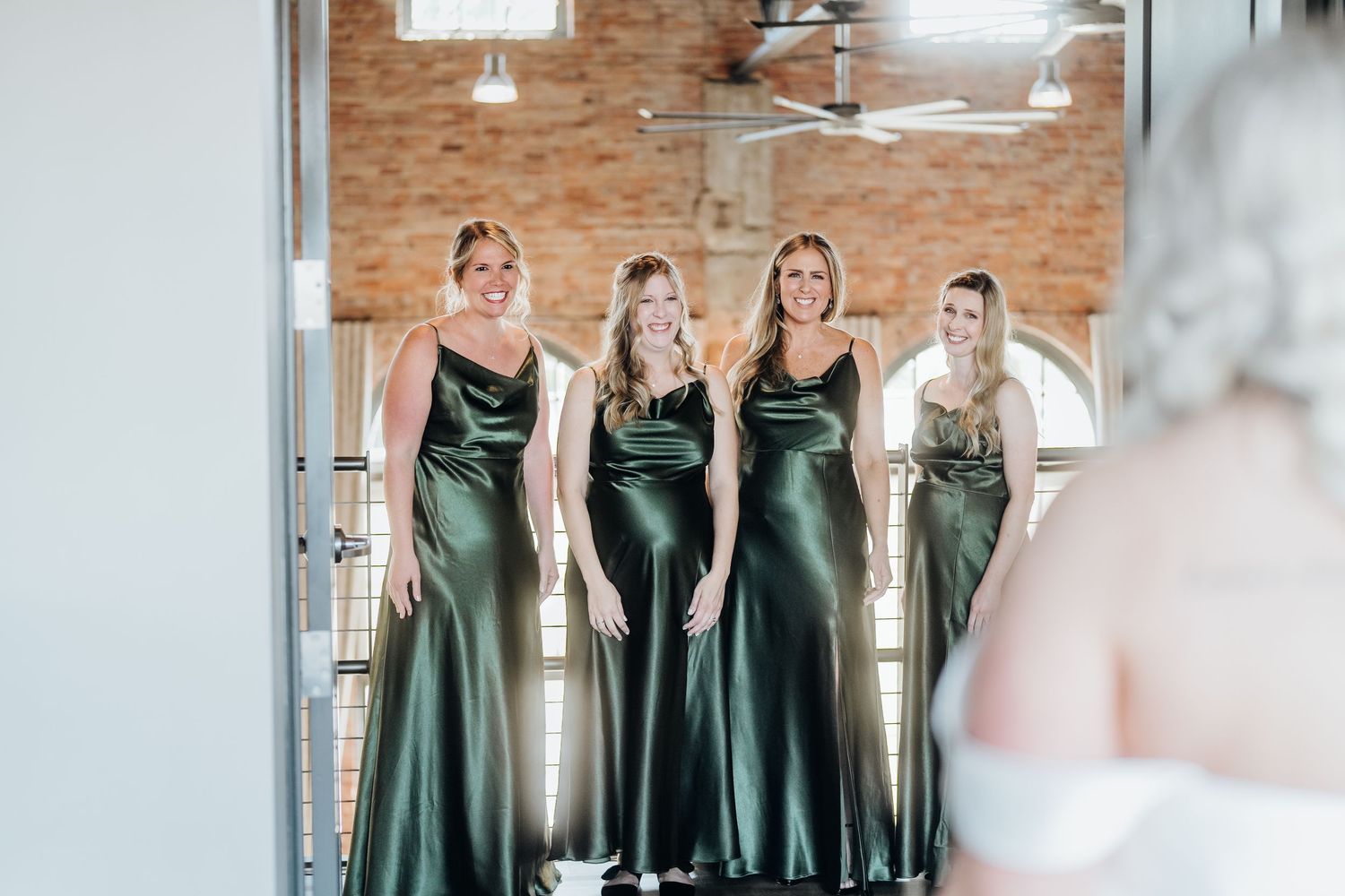 Bridesmaids in emerald green dresses gather for wedding photos in a brick-walled room.