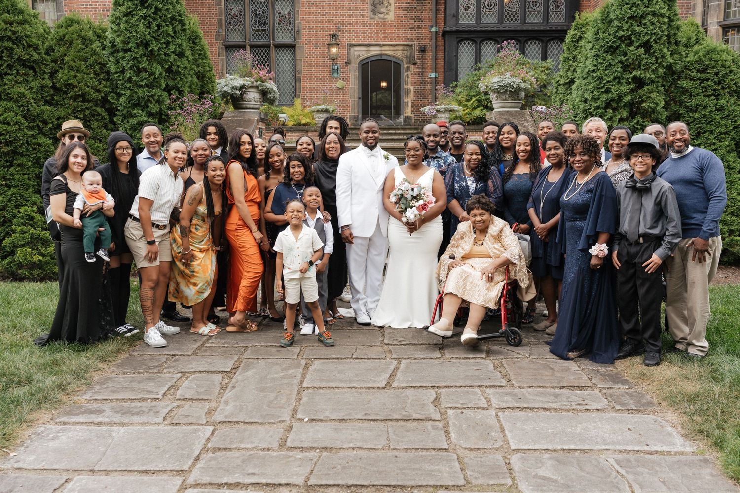 Large wedding party poses together for a group photo outside a brick building with greenery.