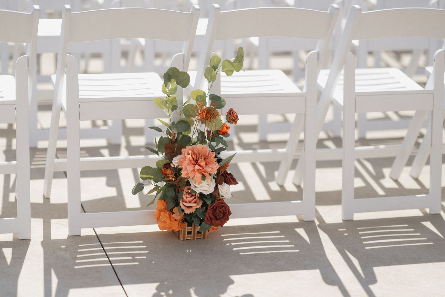 White folding chairs lined up outdoors with coral and peach floral arrangements for a wedding ceremony.