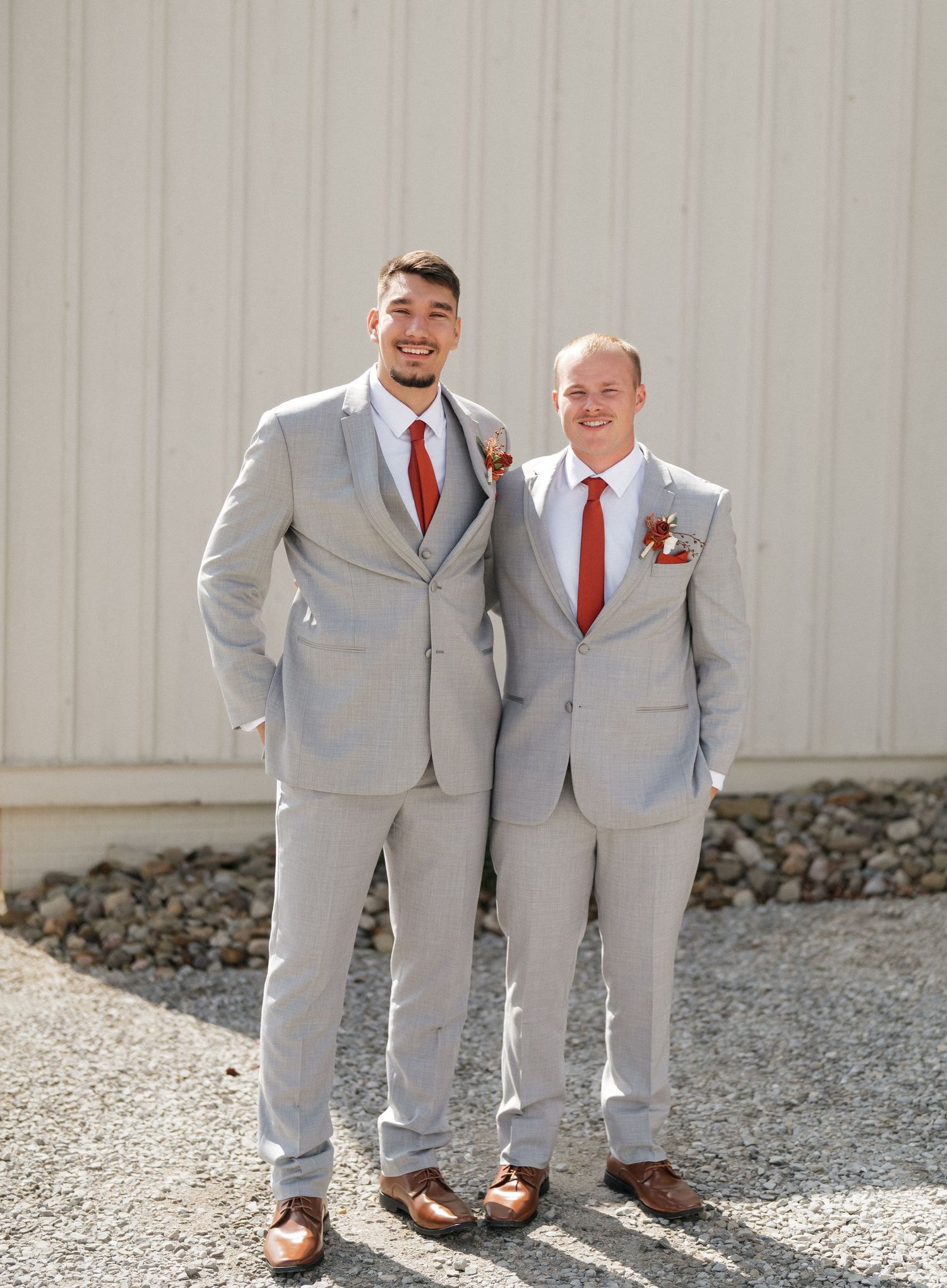 Groomsmen in light grey suits and red ties standing together on gravel.