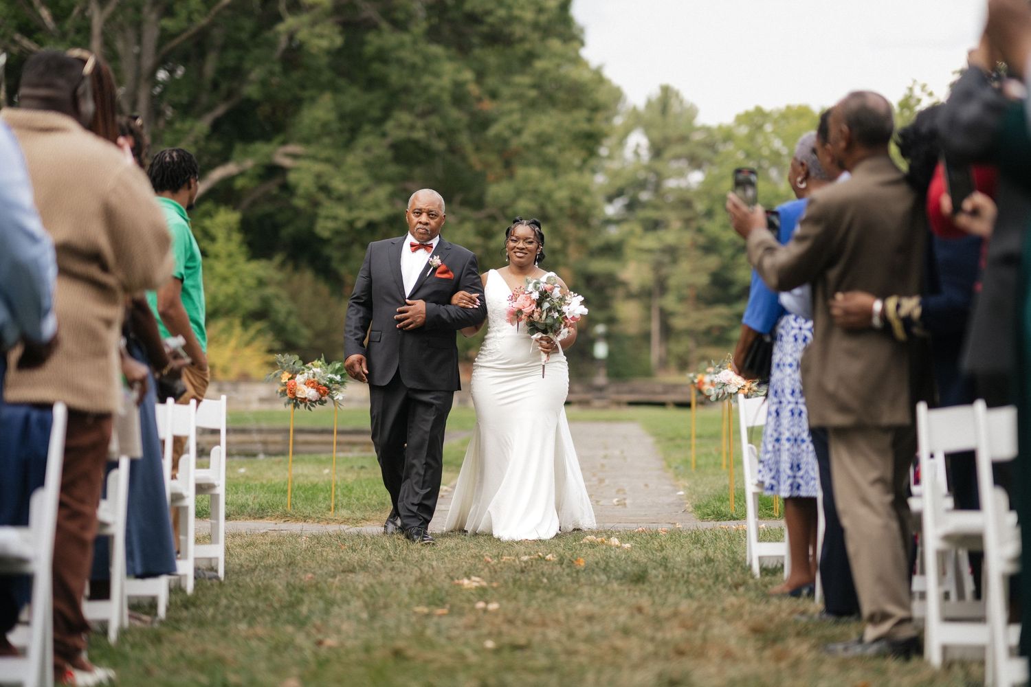 An outdoor wedding ceremony setup with white chairs and guests gathering on a grassy lawn.