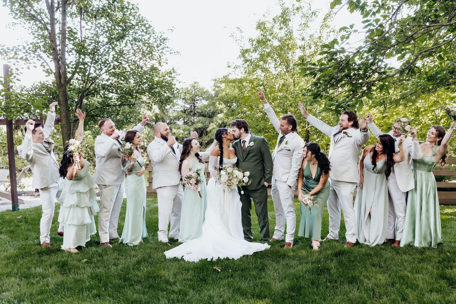 Large wedding party in white and mint green attire poses together on grass under trees.