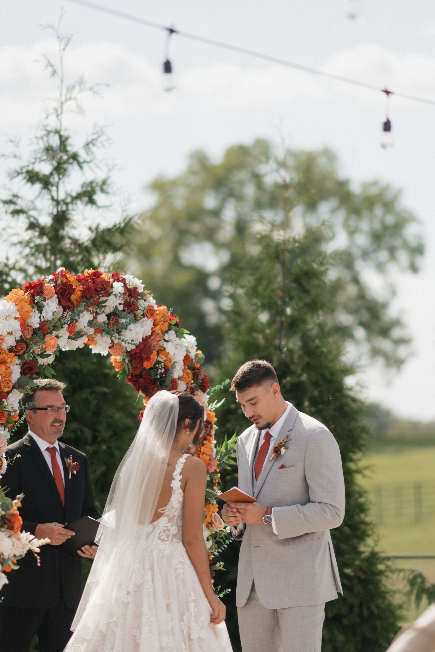 A wedding ceremony under a floral arch with orange and white flowers in an outdoor garden setting.