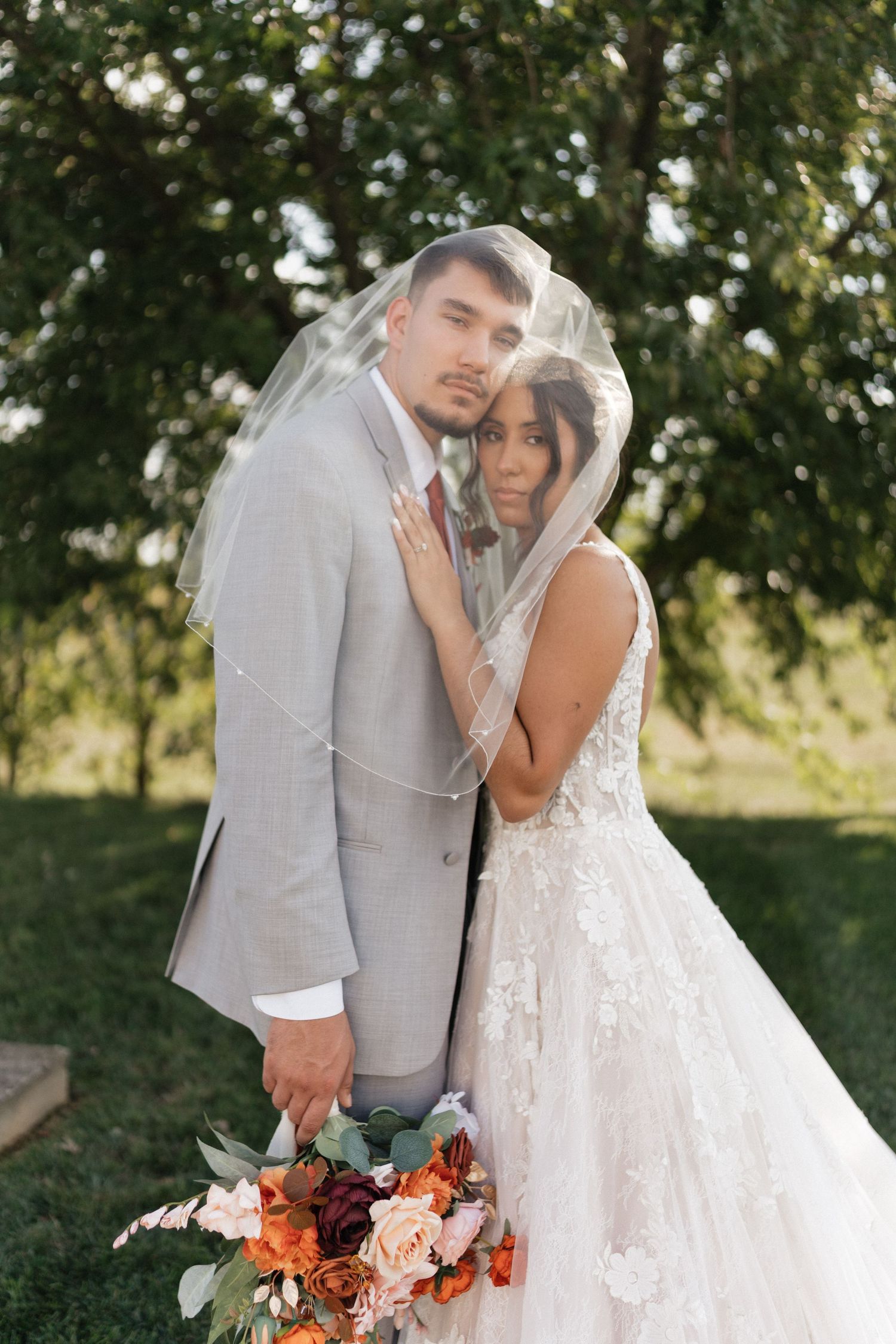 Romantic wedding portrait with couple sharing tender moment under veil with orange and white bouquet.