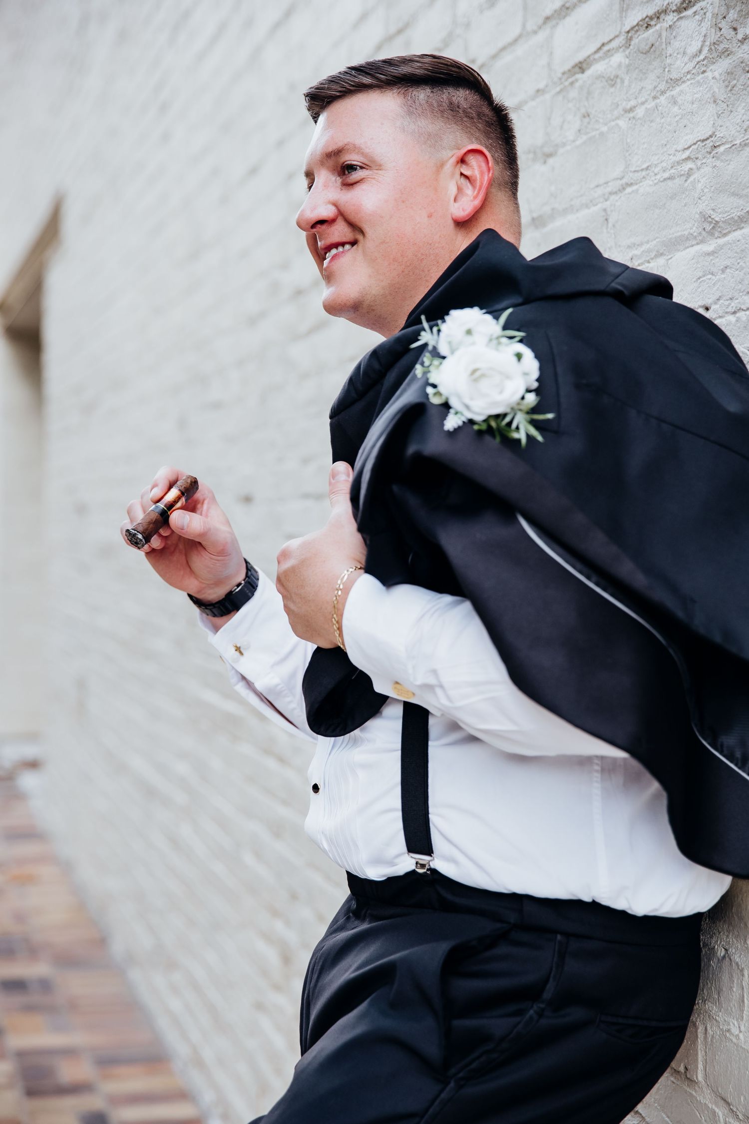 Wedding party member in black tuxedo and white boutonniere leaning against white wall.