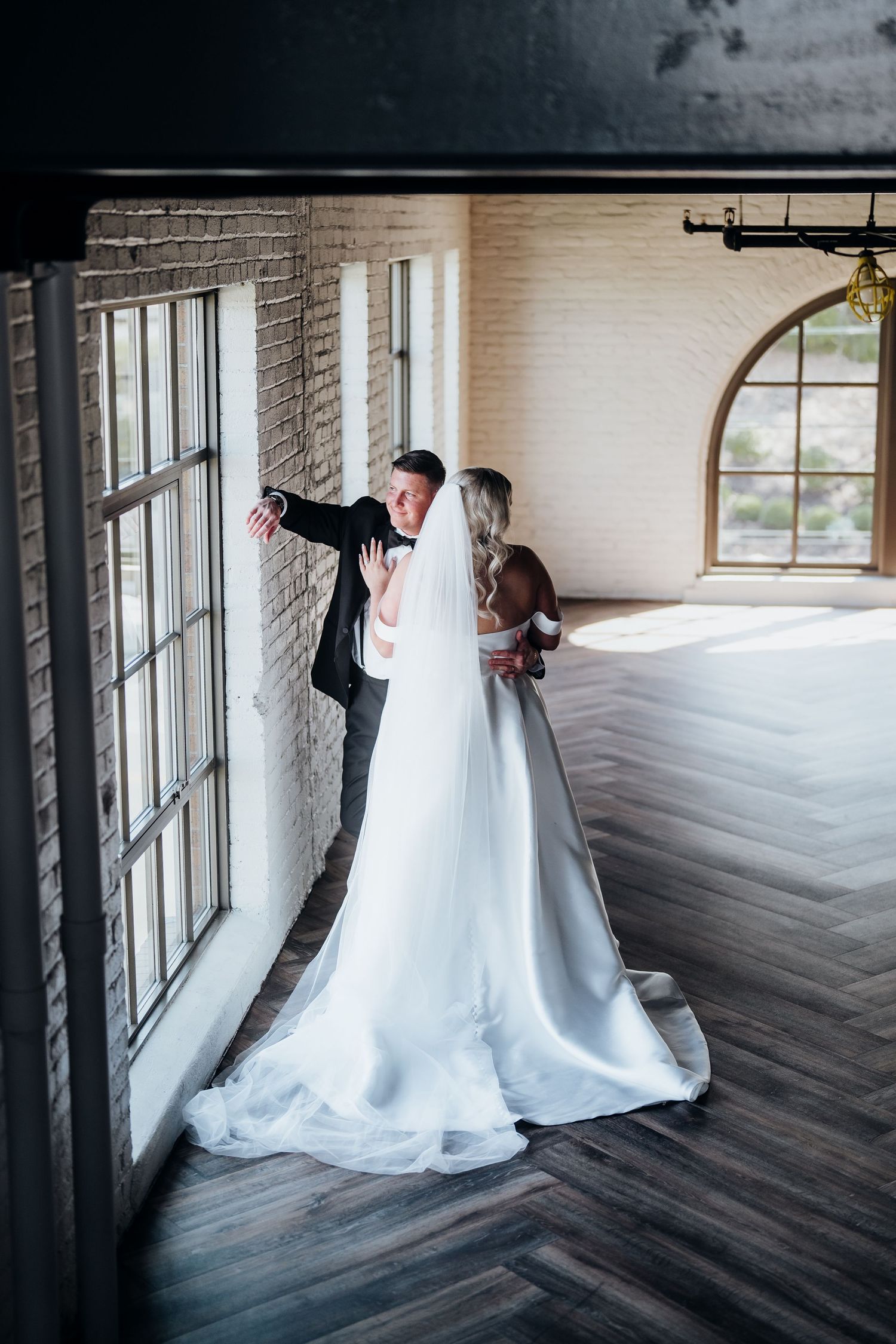 A first look wedding moment captured in a rustic indoor venue with large windows and wooden floors.