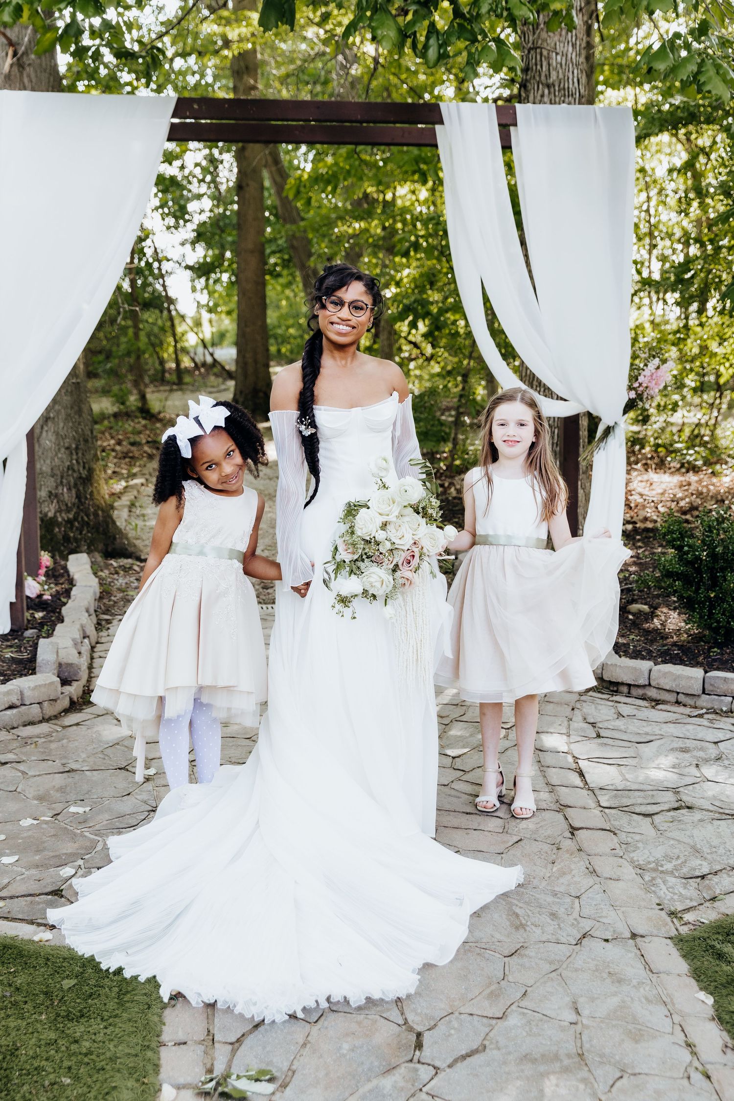 A wedding party poses on stone patio with flowing white dresses and sheer curtains in an outdoor setting.