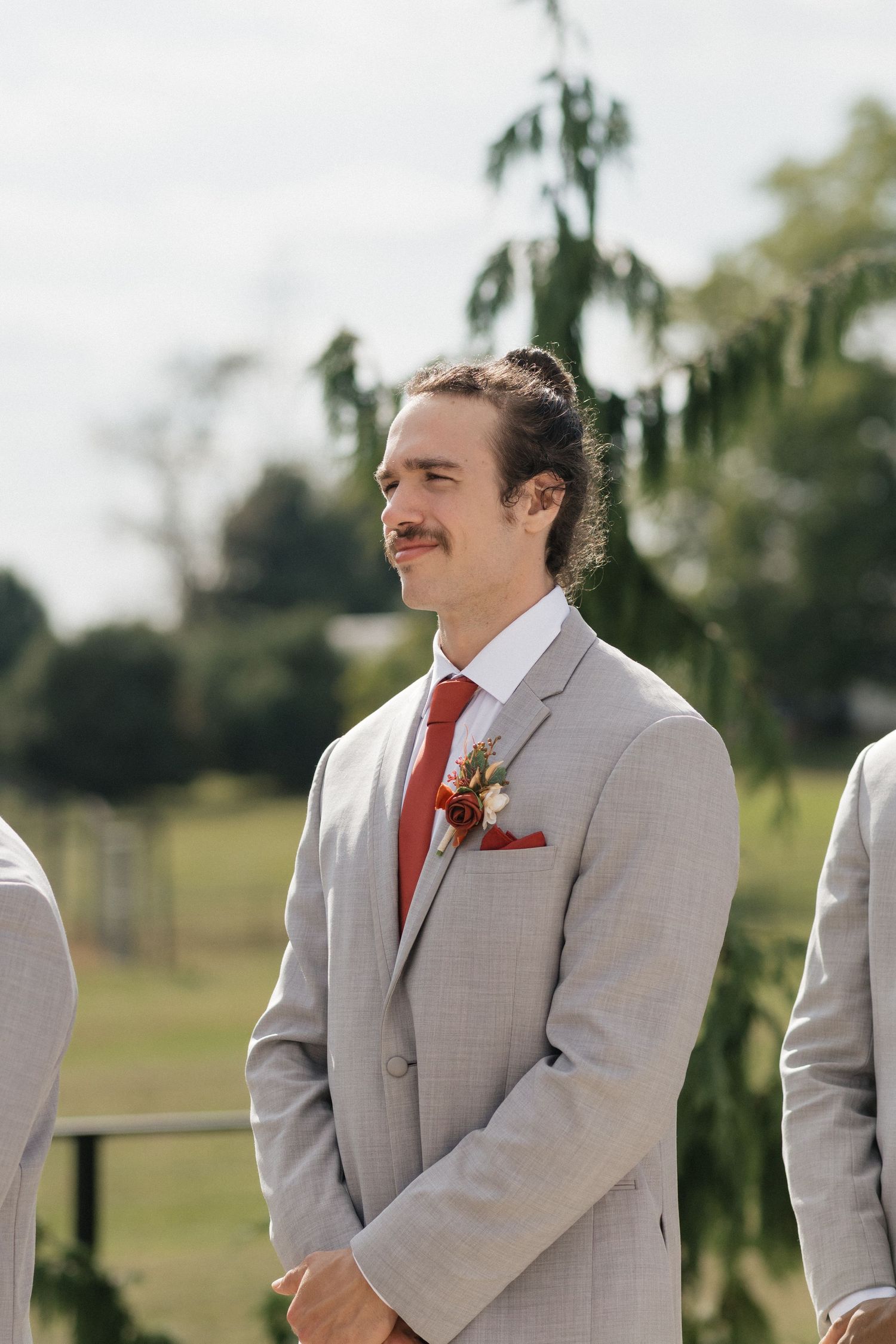 Groomsman in light gray suit with red tie smiling during outdoor wedding ceremony.