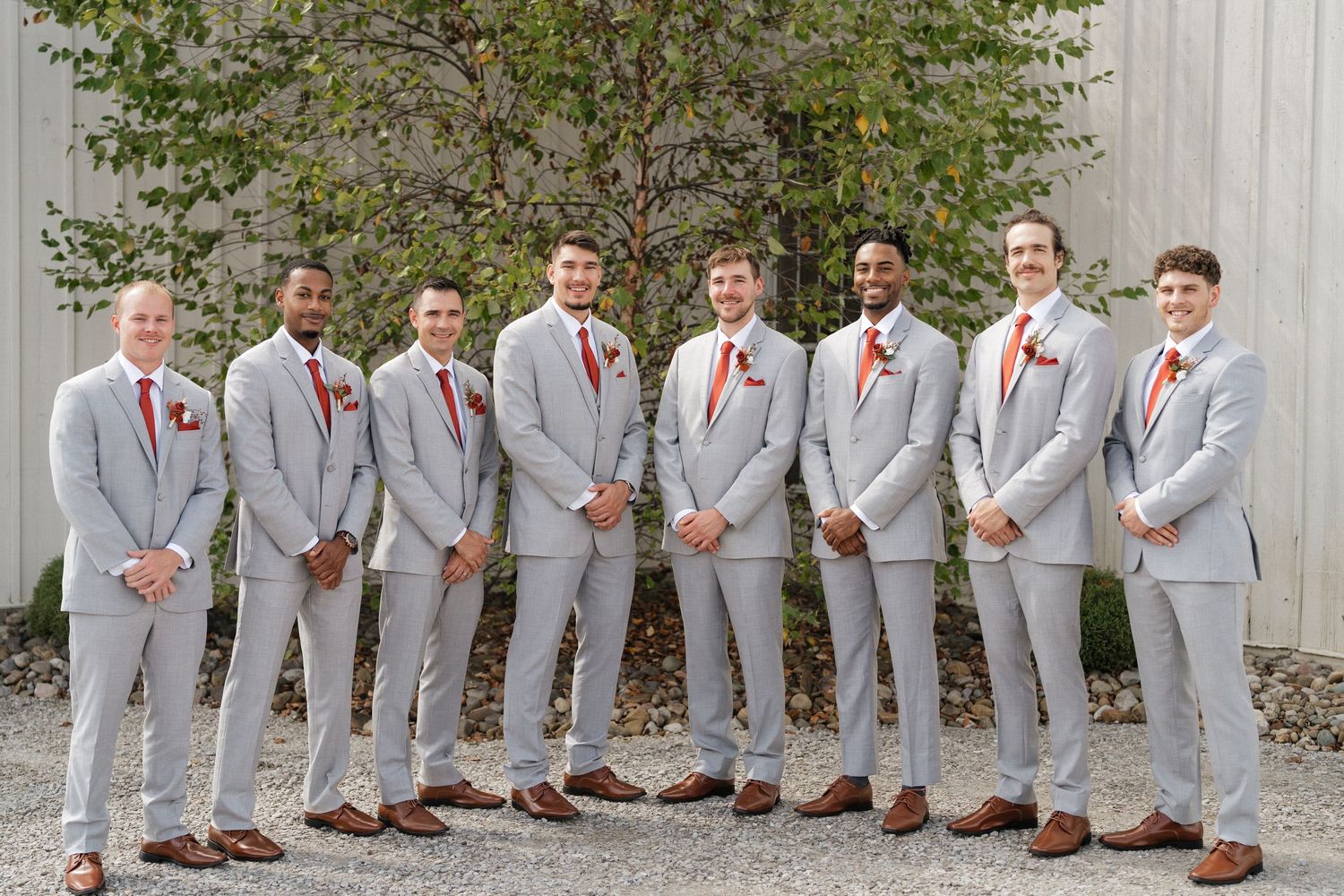 Groomsmen in light grey suits and coral ties lined up against an outdoor wall.