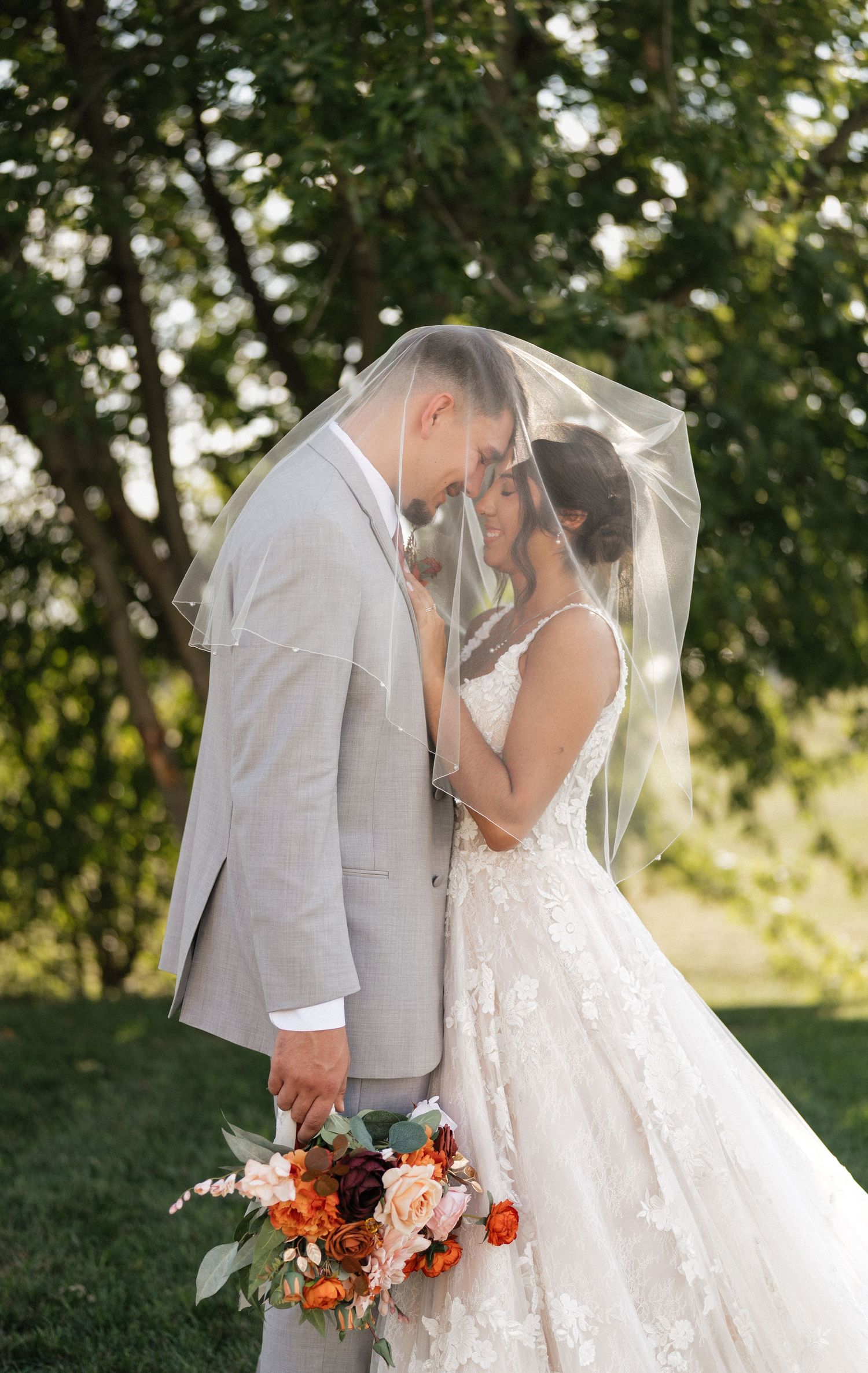 Romantic wedding couple sharing intimate moment under veil with orange floral bouquet.