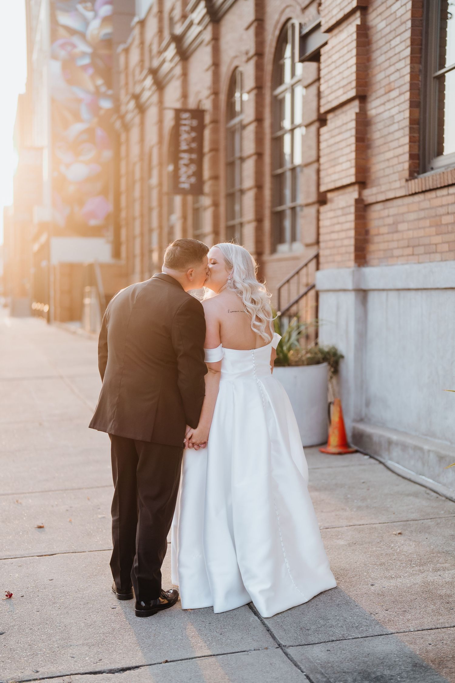 Couple in wedding attire poses outside a historic brick building during golden hour.