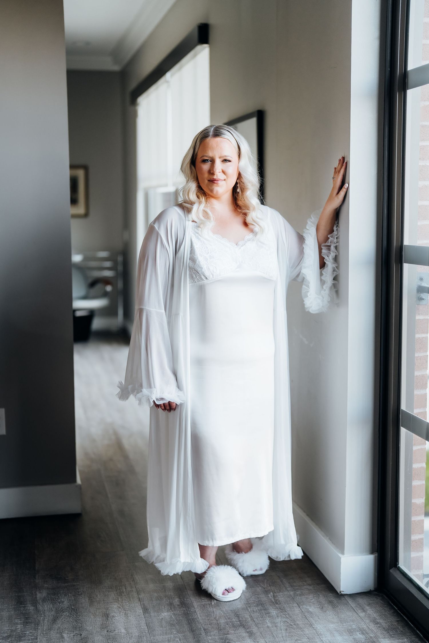 Person in a white flowing dress standing near large windows in a modern interior.