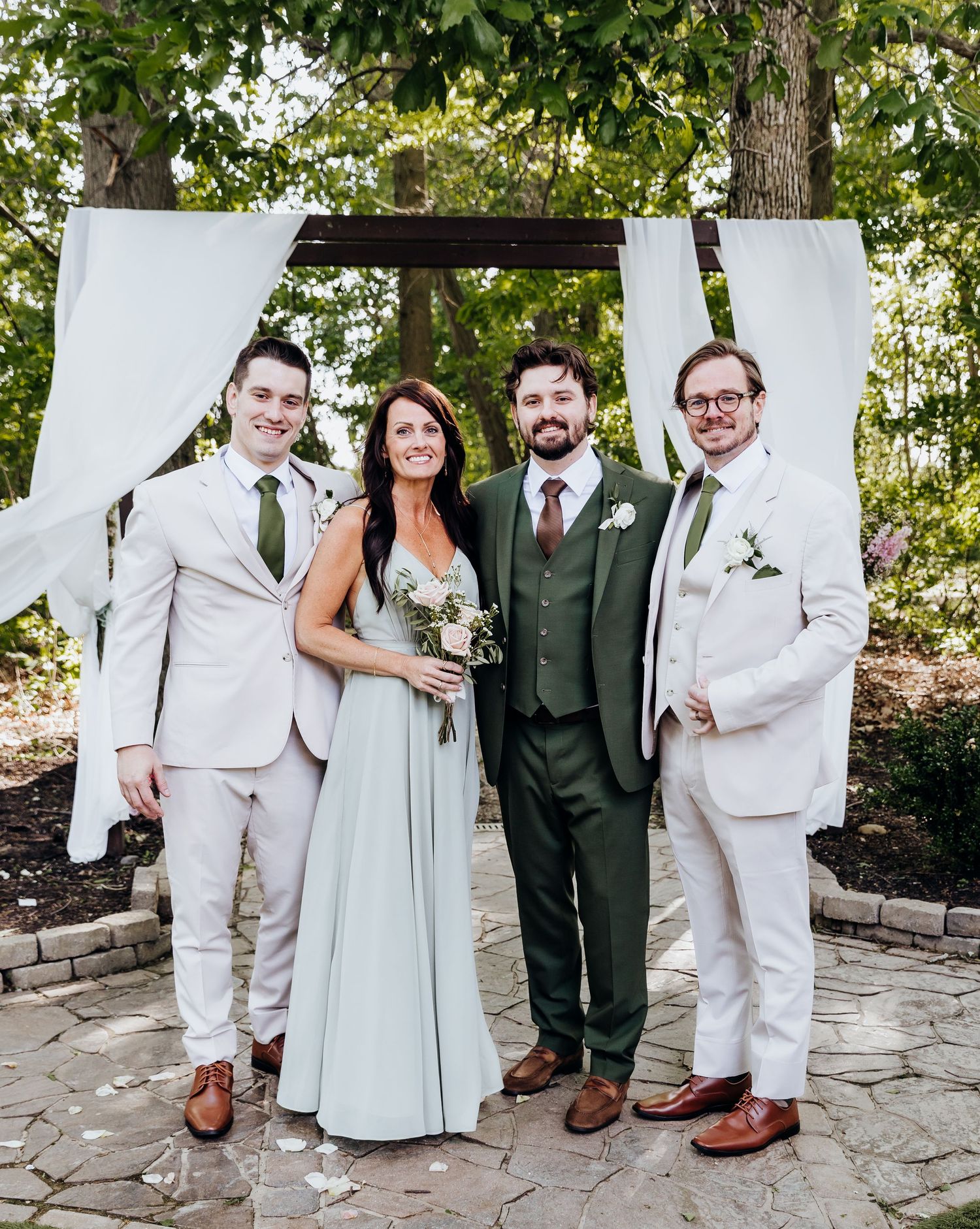 Wedding party poses under white draped arch with groomsmen in light suits and dark green accent.