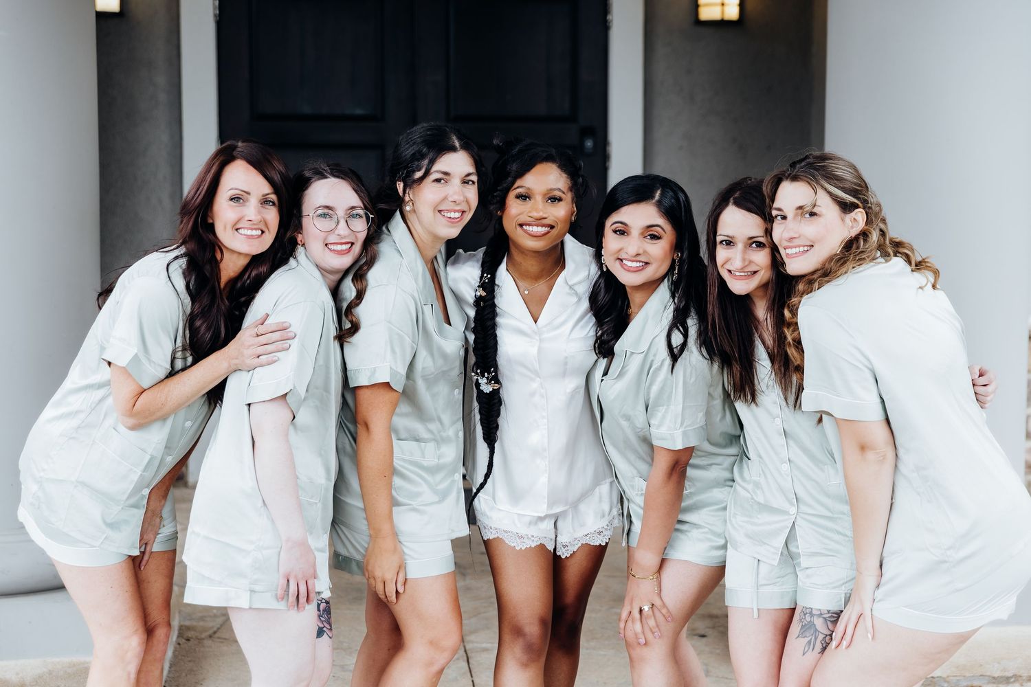 Group of bridesmaids in white robes laughing together on a columned porch.