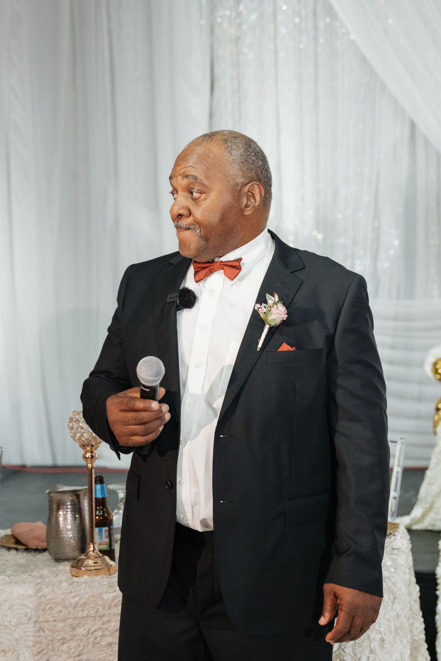 A gentleman in a classic black tuxedo and red bowtie smiles during wedding reception festivities.