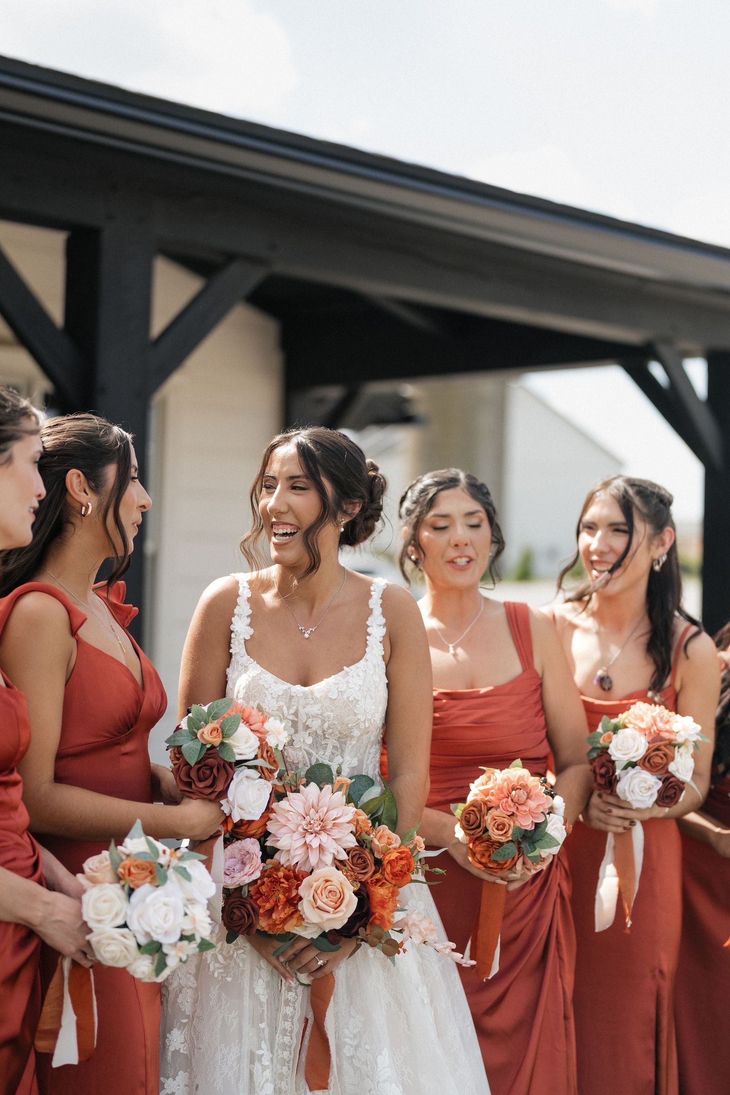 Wedding party in burgundy bridesmaid dresses and white bridal gown holding floral bouquets at outdoor venue.