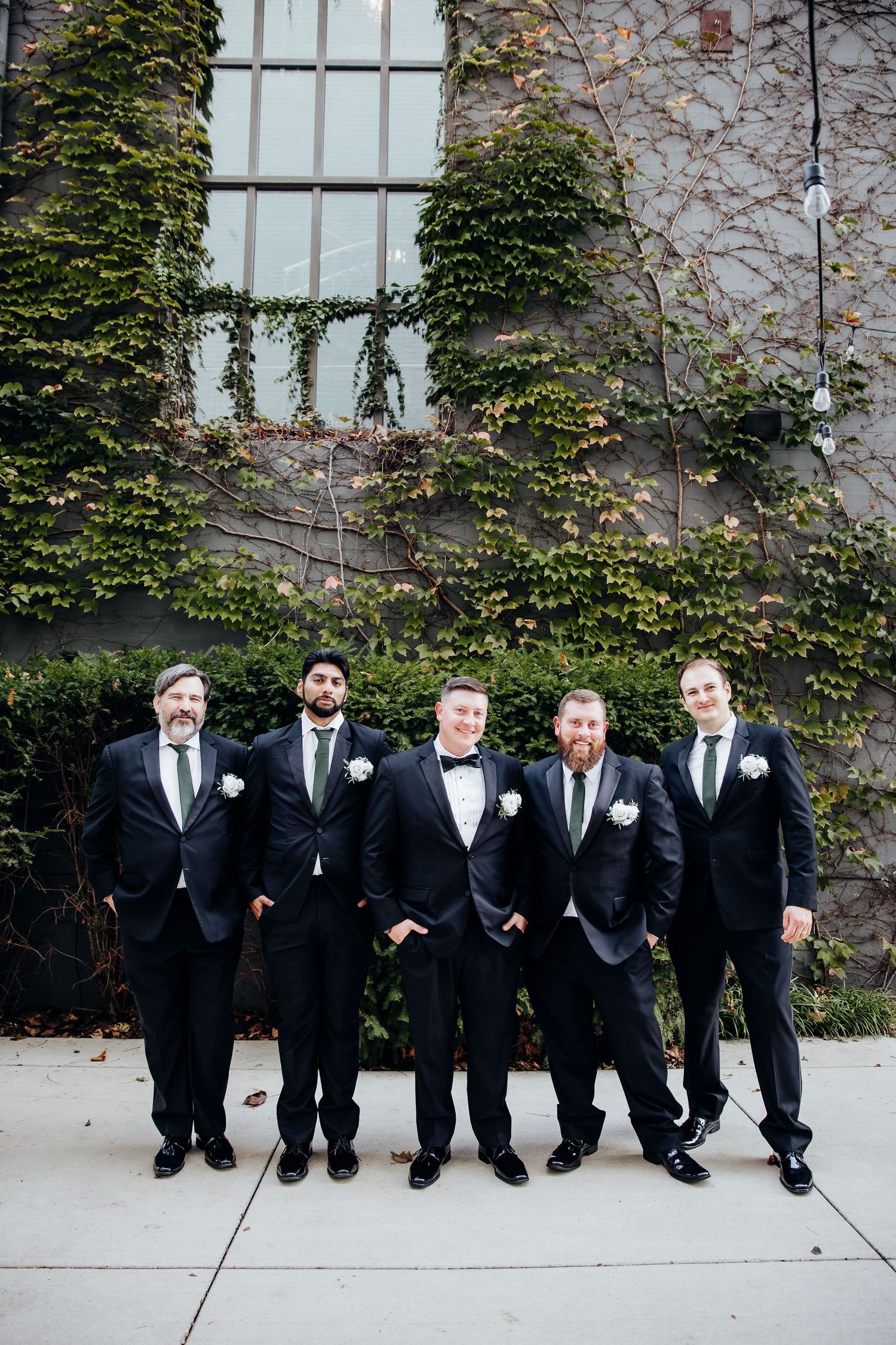 Groomsmen in black tuxedos pose casually sitting against an ivy-covered wall.
