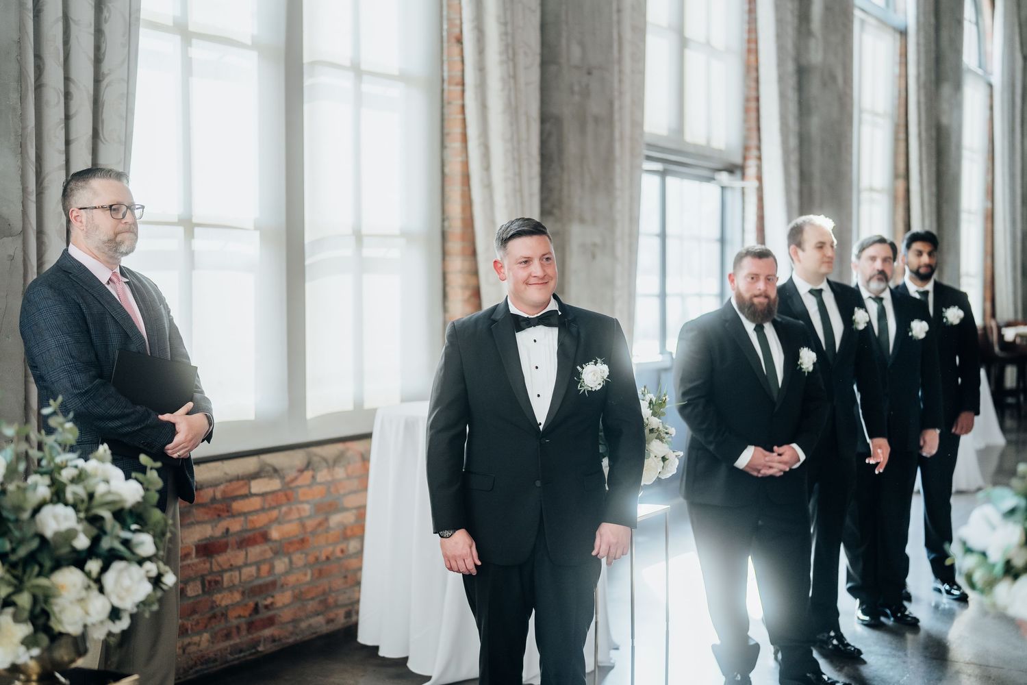 A wedding party of groomsmen in black tuxedos line up in an industrial-chic venue with tall windows.