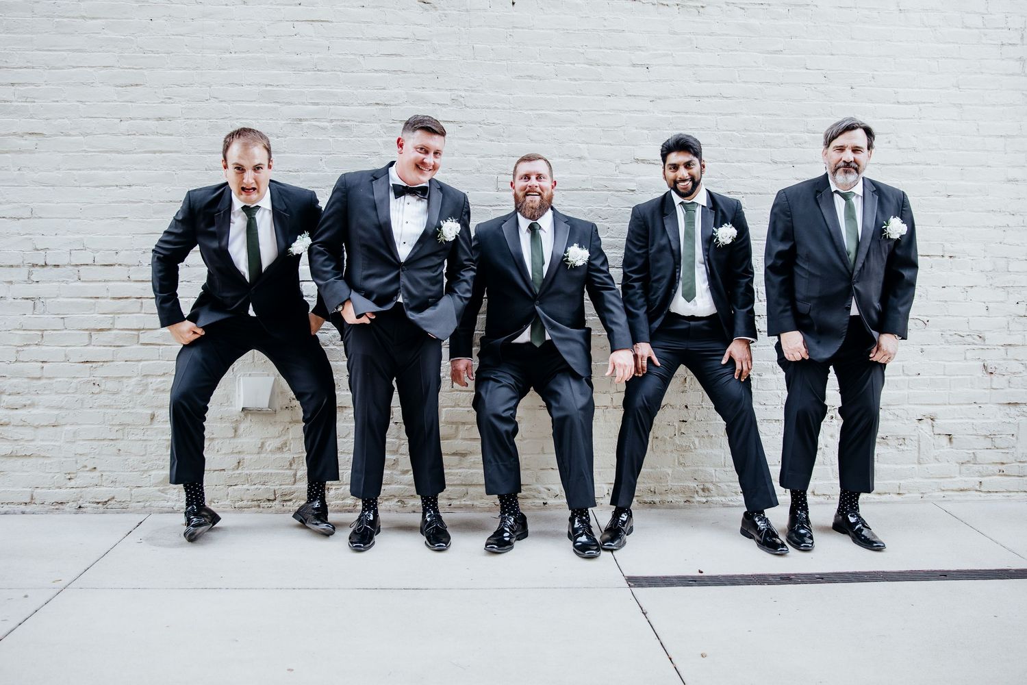 Groomsmen in dark suits sit casually on white brick wall for wedding party portrait.