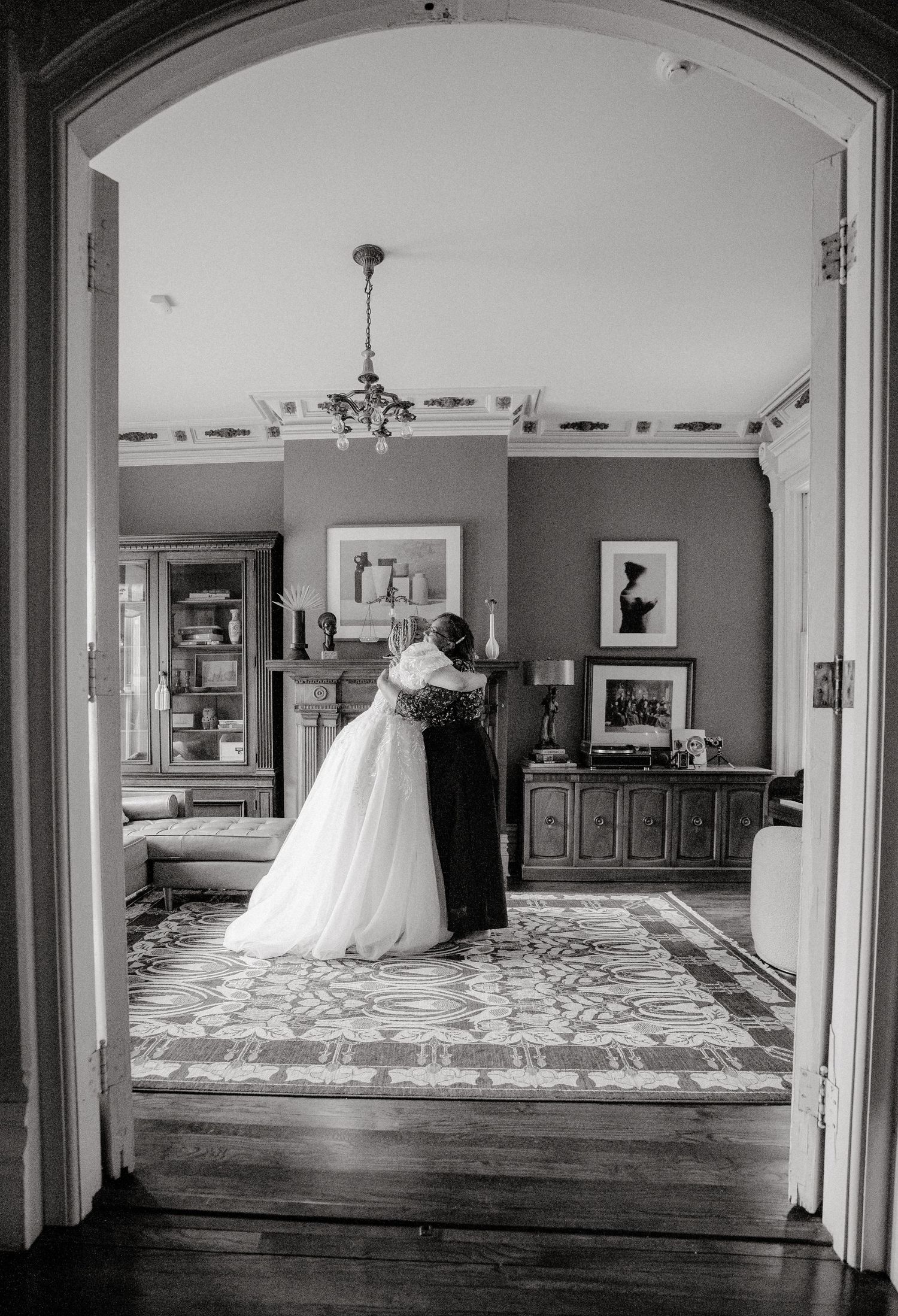 A black and white wedding photo captures a couple embracing in an elegant room framed by an archway.