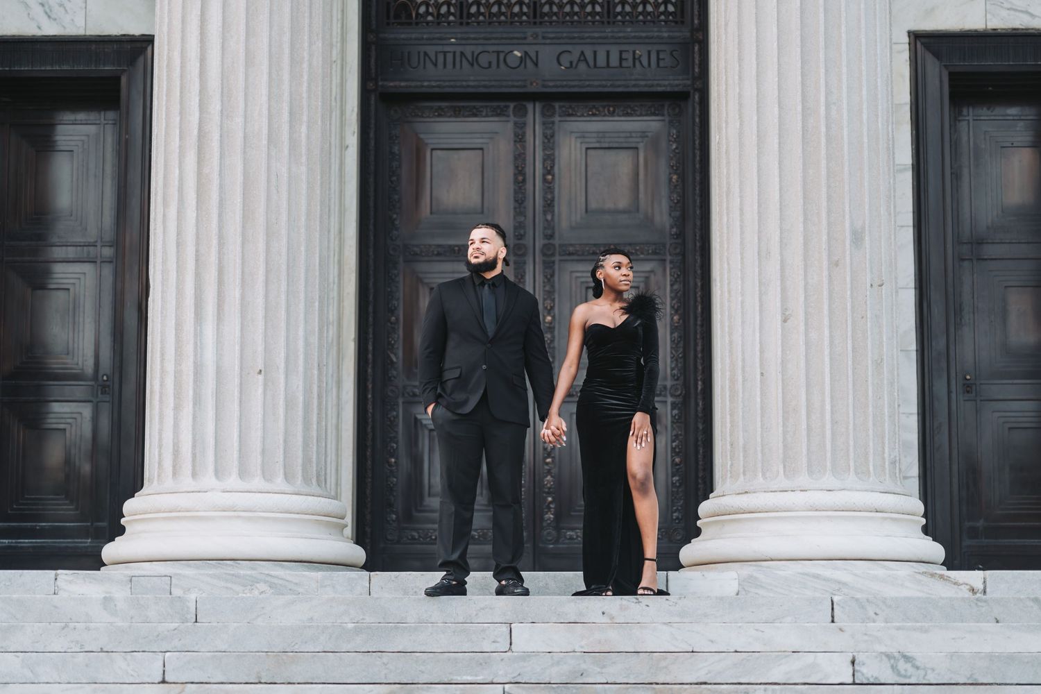 A couple poses dramatically between classical columns in formal black attire.