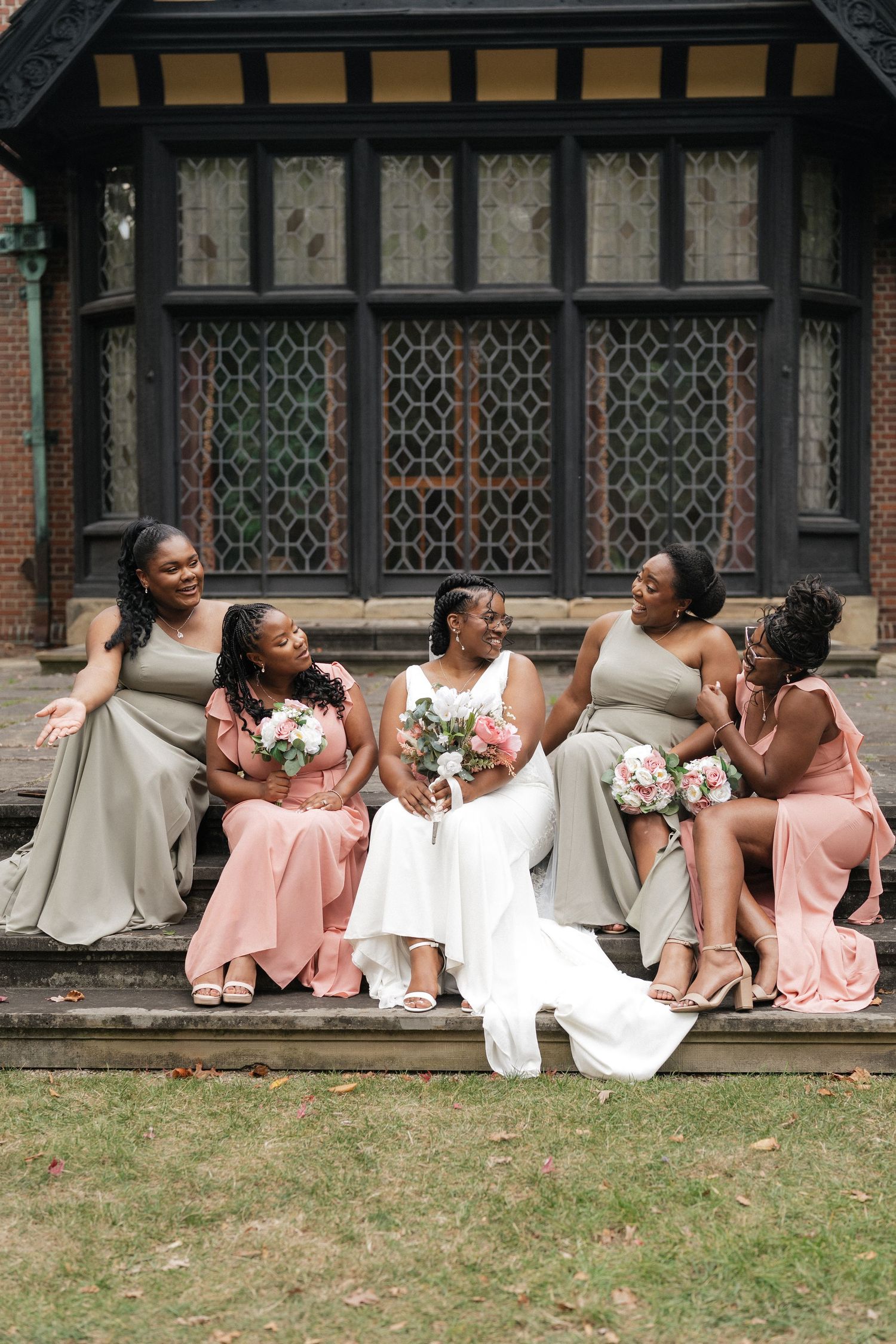 Wedding party relaxes on grass in front of Tudor-style building with ornate windows.