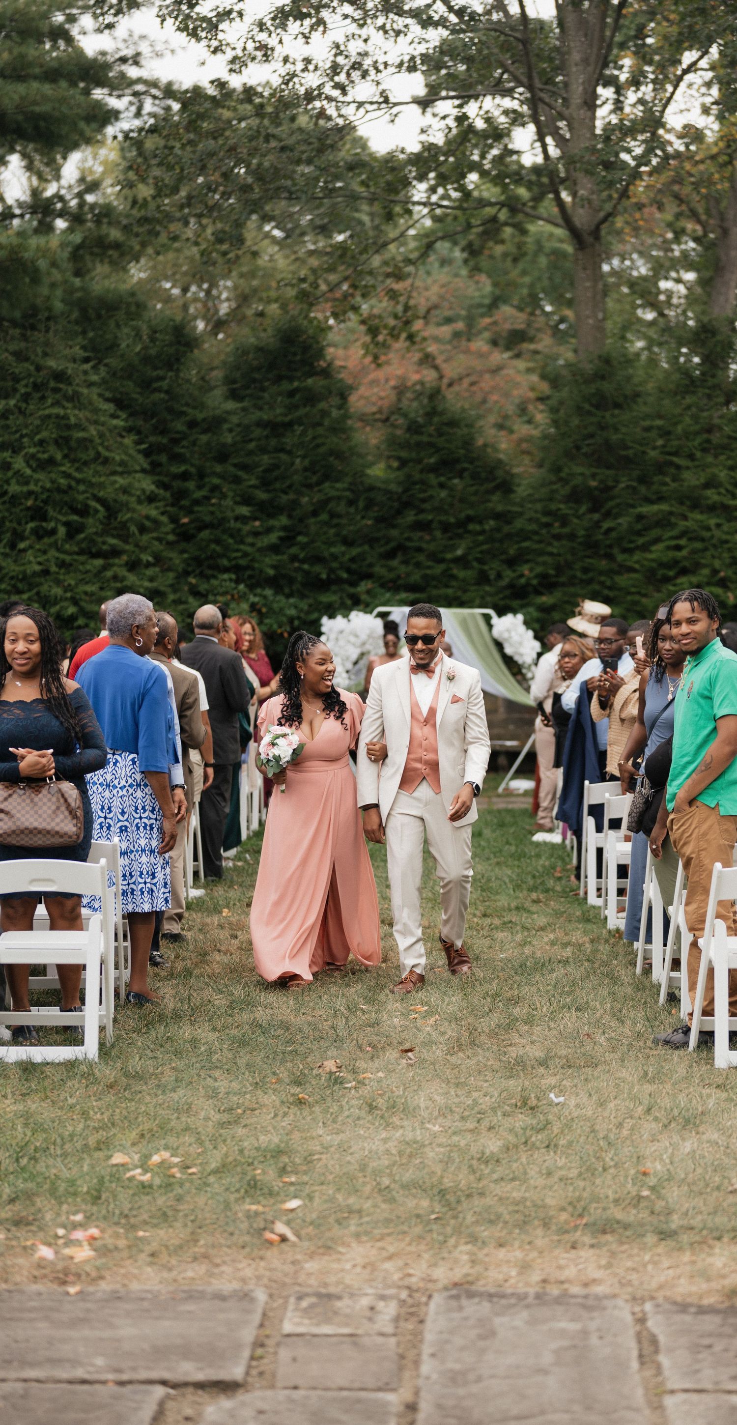 Wedding guests line white chairs as a couple walks down the aisle at an outdoor garden ceremony.