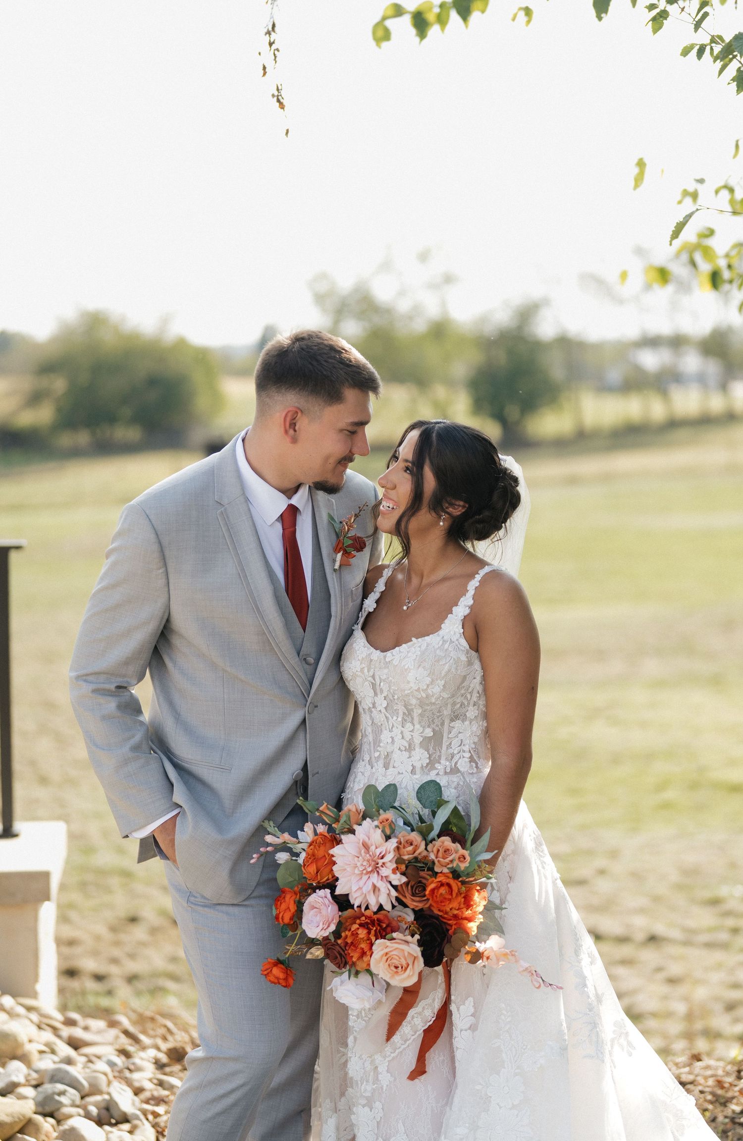Romantic wedding portrait with couple holding coral and peach floral bouquet in outdoor setting.