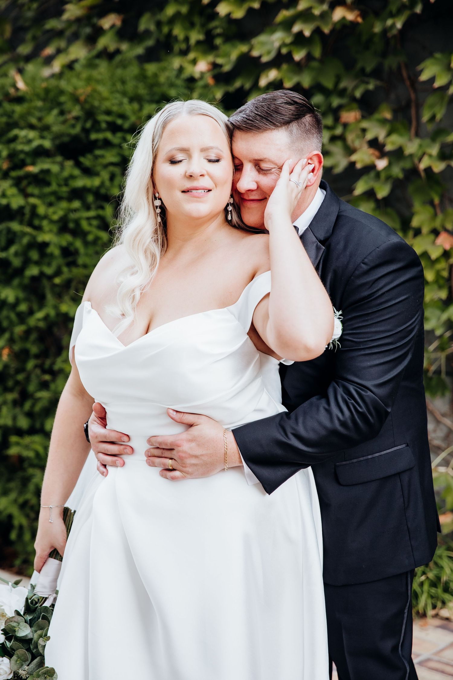 Intimate wedding couple moment captured against lush garden greenery backdrop.