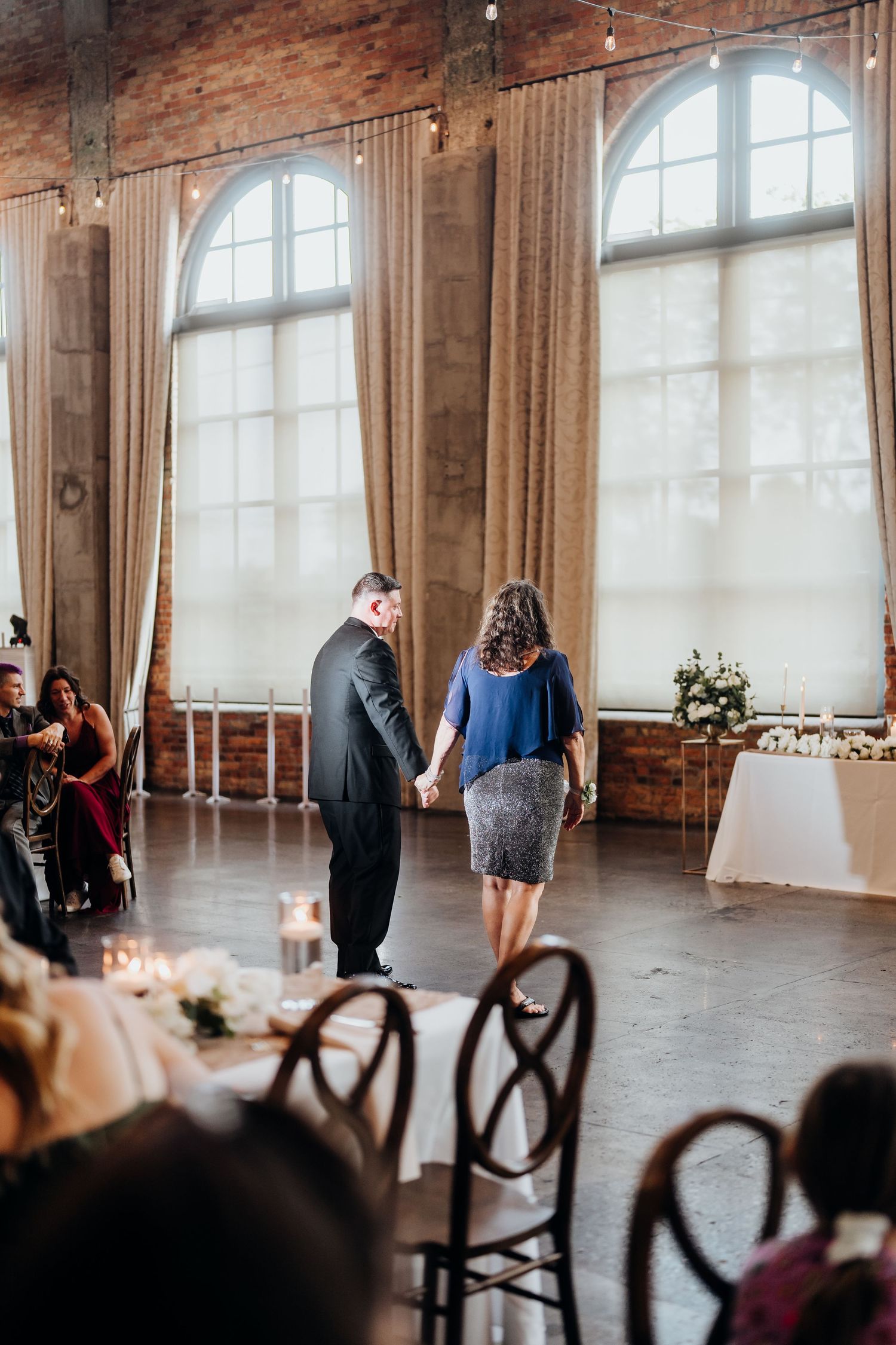 Wedding guests mingle in an elegant venue with arched windows and exposed brick walls.