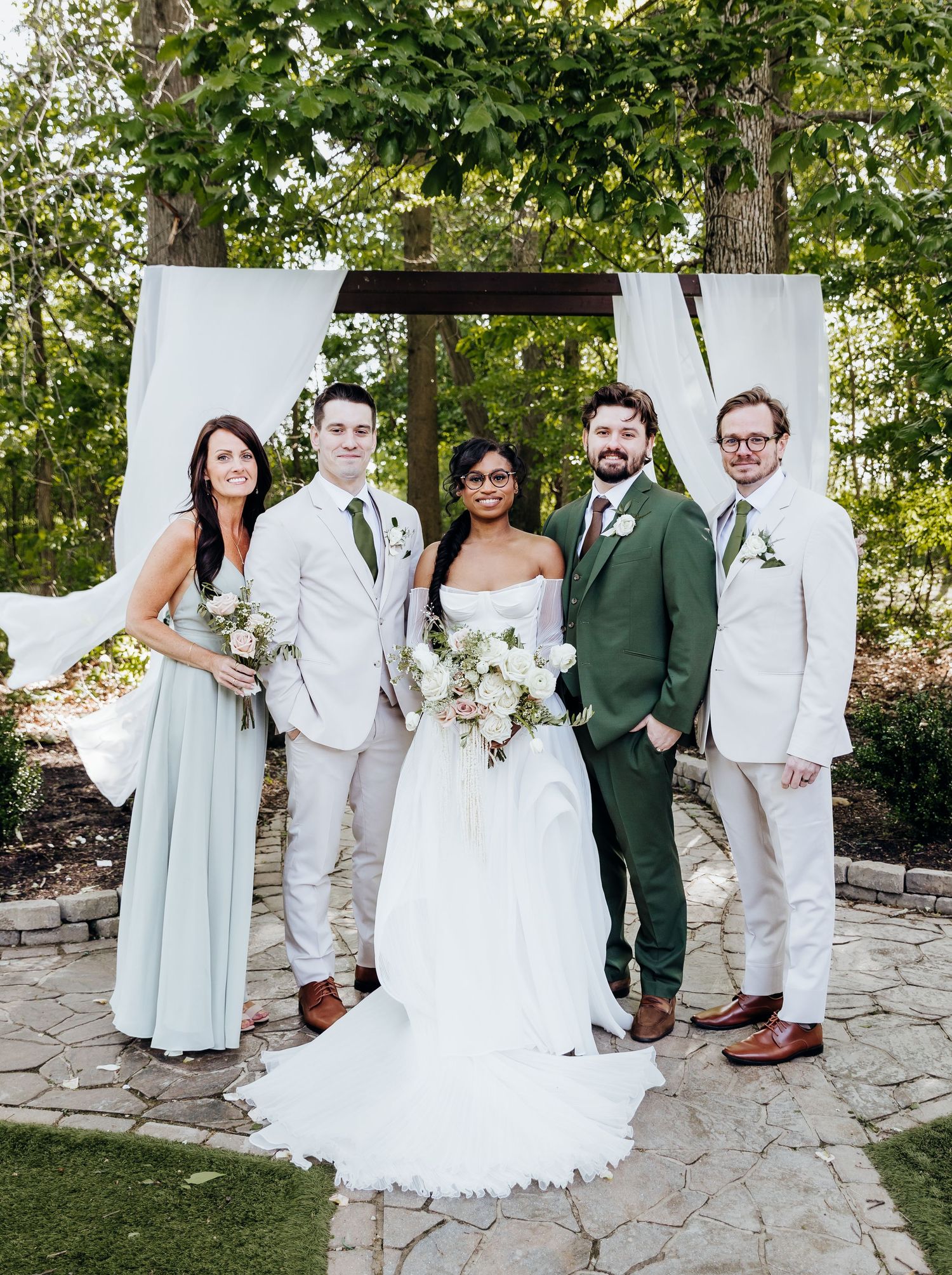 A wedding party poses outdoors under a white draped arch with greenery, featuring light and dark colored attire.