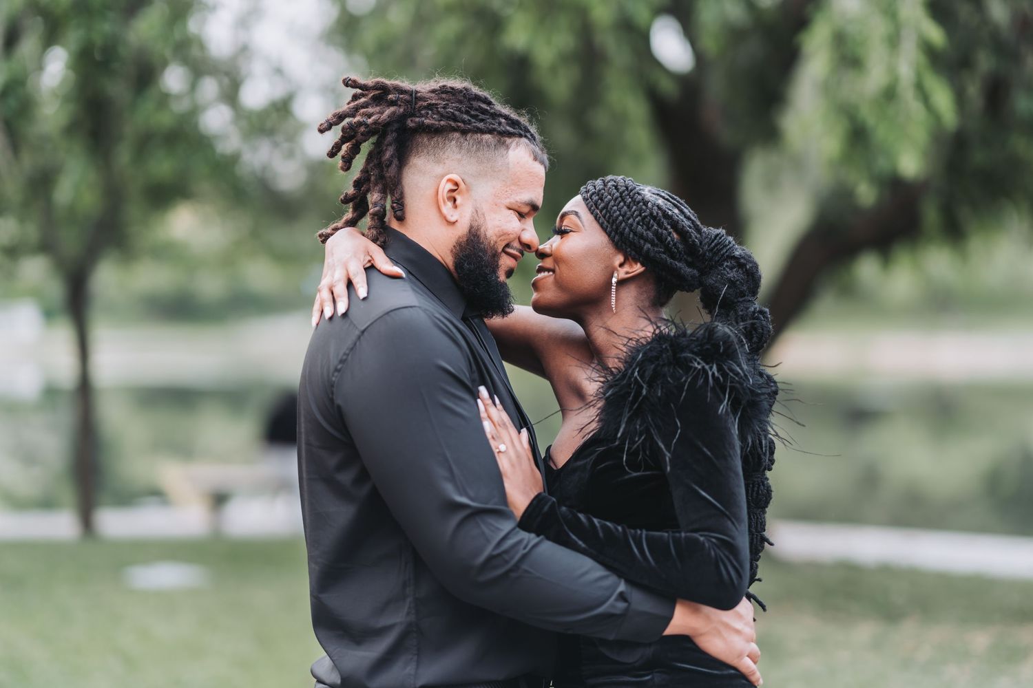 A couple embraces outdoors near a lake, sharing a romantic moment in coordinated dark formal wear.