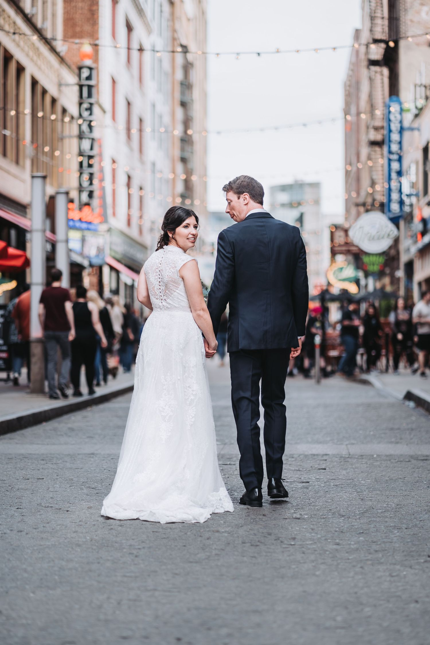 A couple walks down a bustling city street after their wedding, holding hands and looking back.