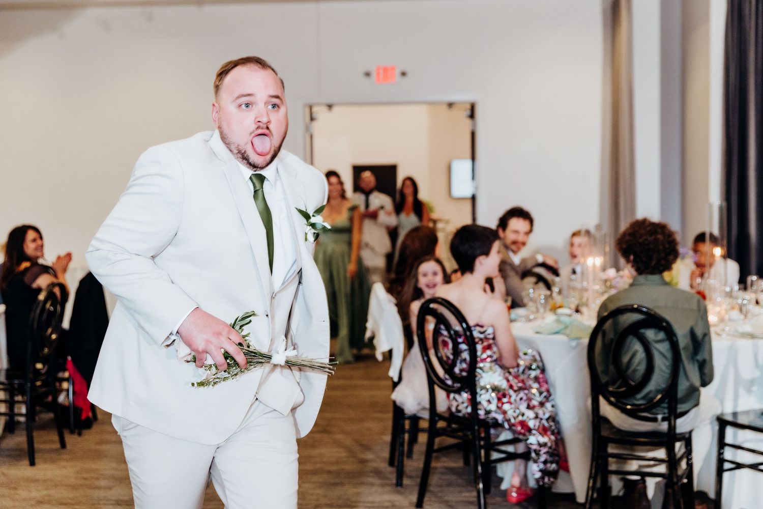 A person in a white suit shows excitement during a wedding reception in an indoor venue.